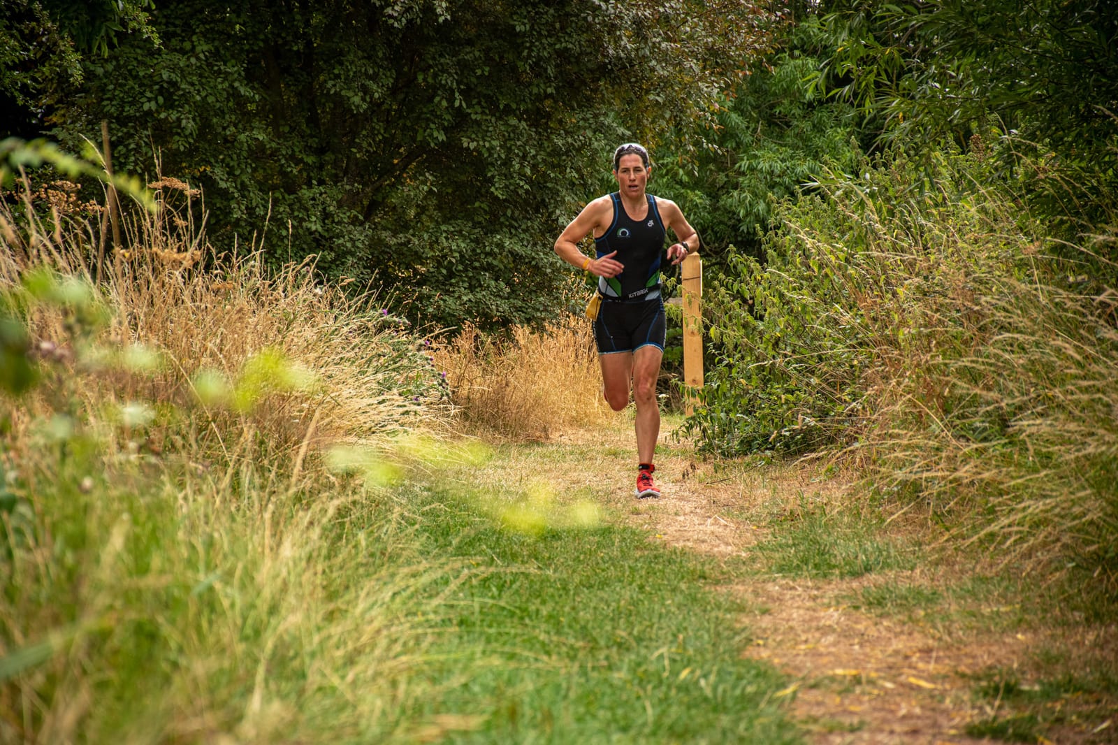 A person wearing athletic gear and a backwards cap runs on a dirt trail surrounded by tall grass and trees, with greenery and foliage in the background.