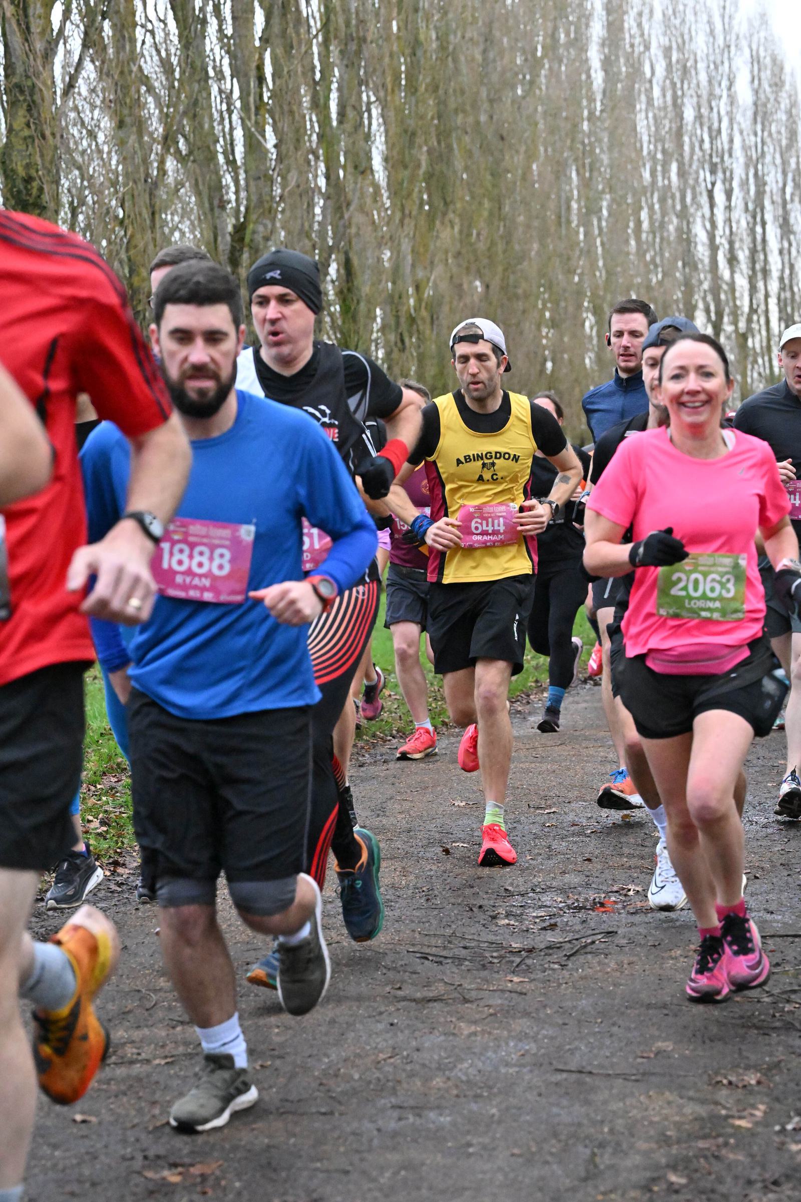 A group of runners participating in a race on a tree-lined path. They are wearing various athletic outfits, some with numbered bibs. The atmosphere appears energetic and focused as they move forward in unison.