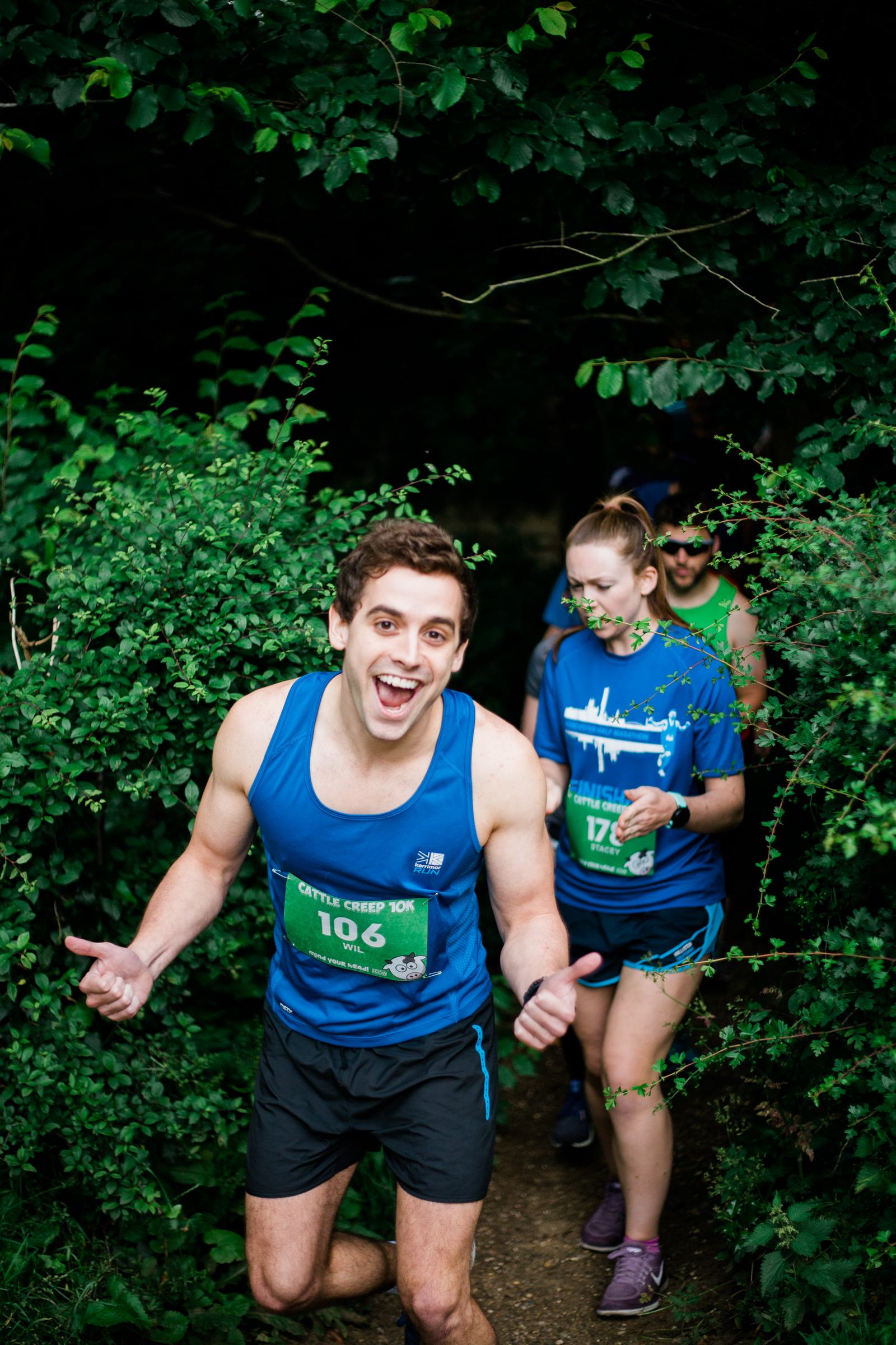 A group of people in athletic gear running on a narrow, leafy trail. The leading man, wearing a blue tank top and smiling, is giving a thumbs-up. Two others follow closely, surrounded by greenery. They all have race bibs pinned to their shirts.