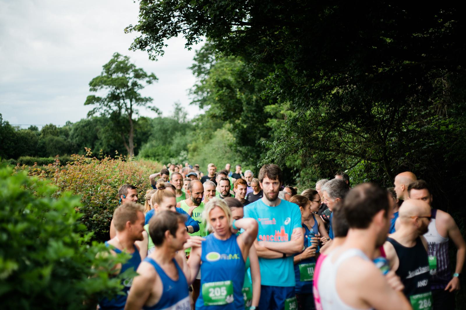 A group of people in athletic gear stand together outdoors, surrounded by trees and greenery. They appear to be participating in a race or marathon, with some individuals wearing bibs. The atmosphere is lively and focused.