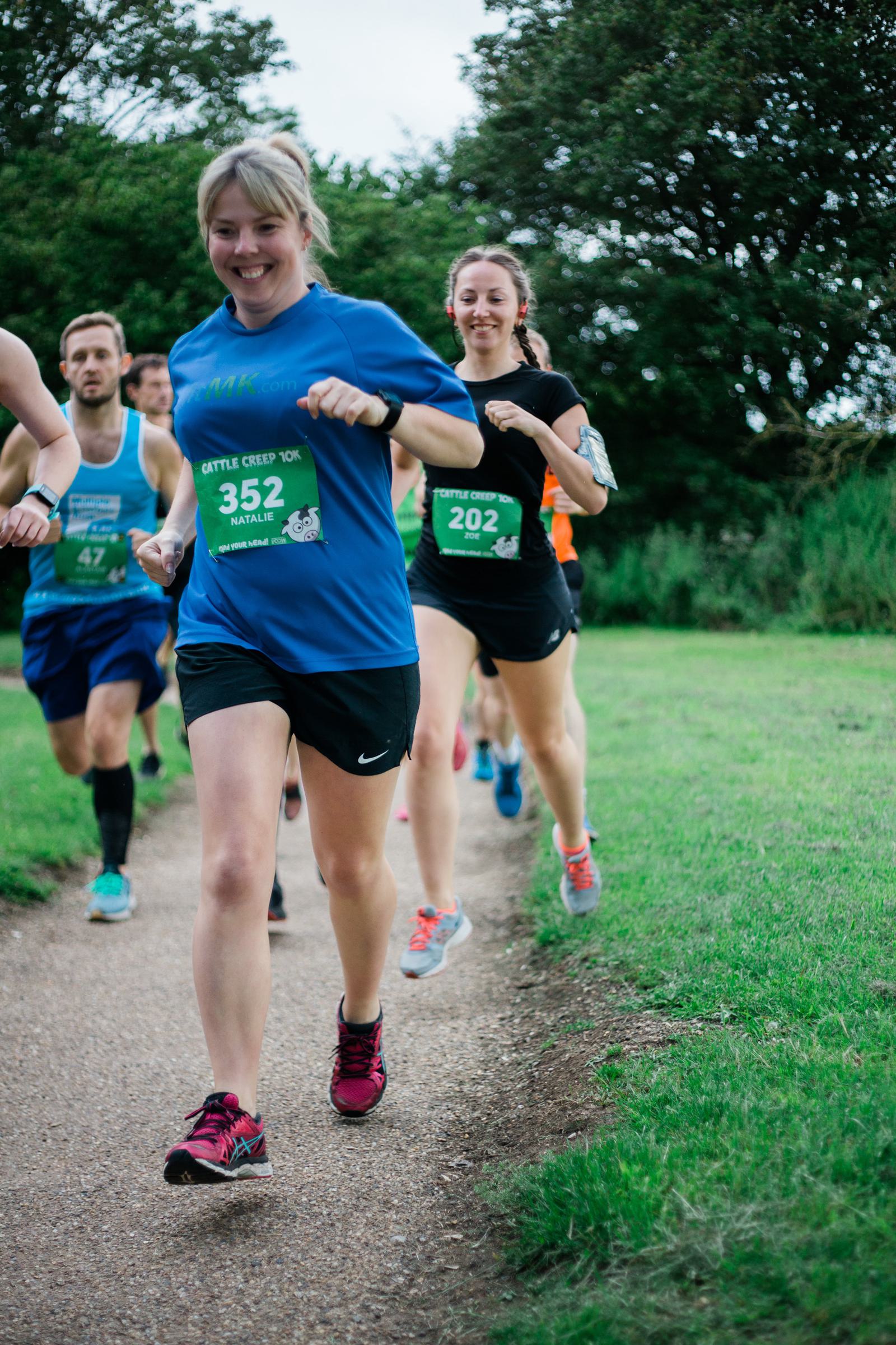 A group of runners, including a woman in a blue shirt, participate in a race on a gravel path in a park. They wear numbered bibs and run energetically. The background features green grass and trees.