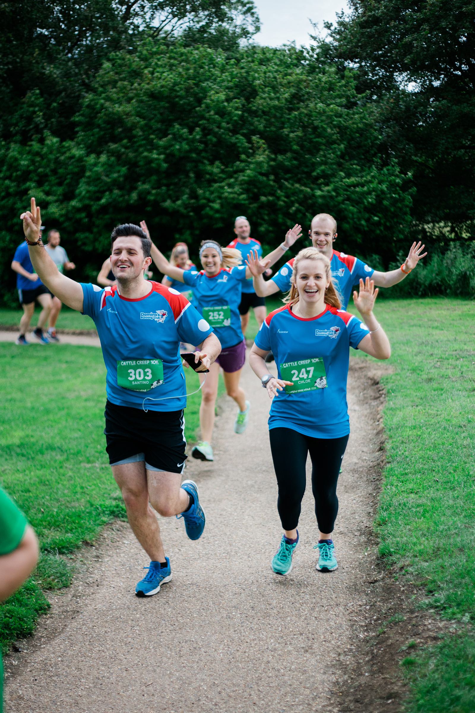 People in blue uniforms are joyfully participating in an outdoor run on a gravel path, surrounded by greenery. They wave towards the camera while wearing race bibs. Some trees are visible in the background.