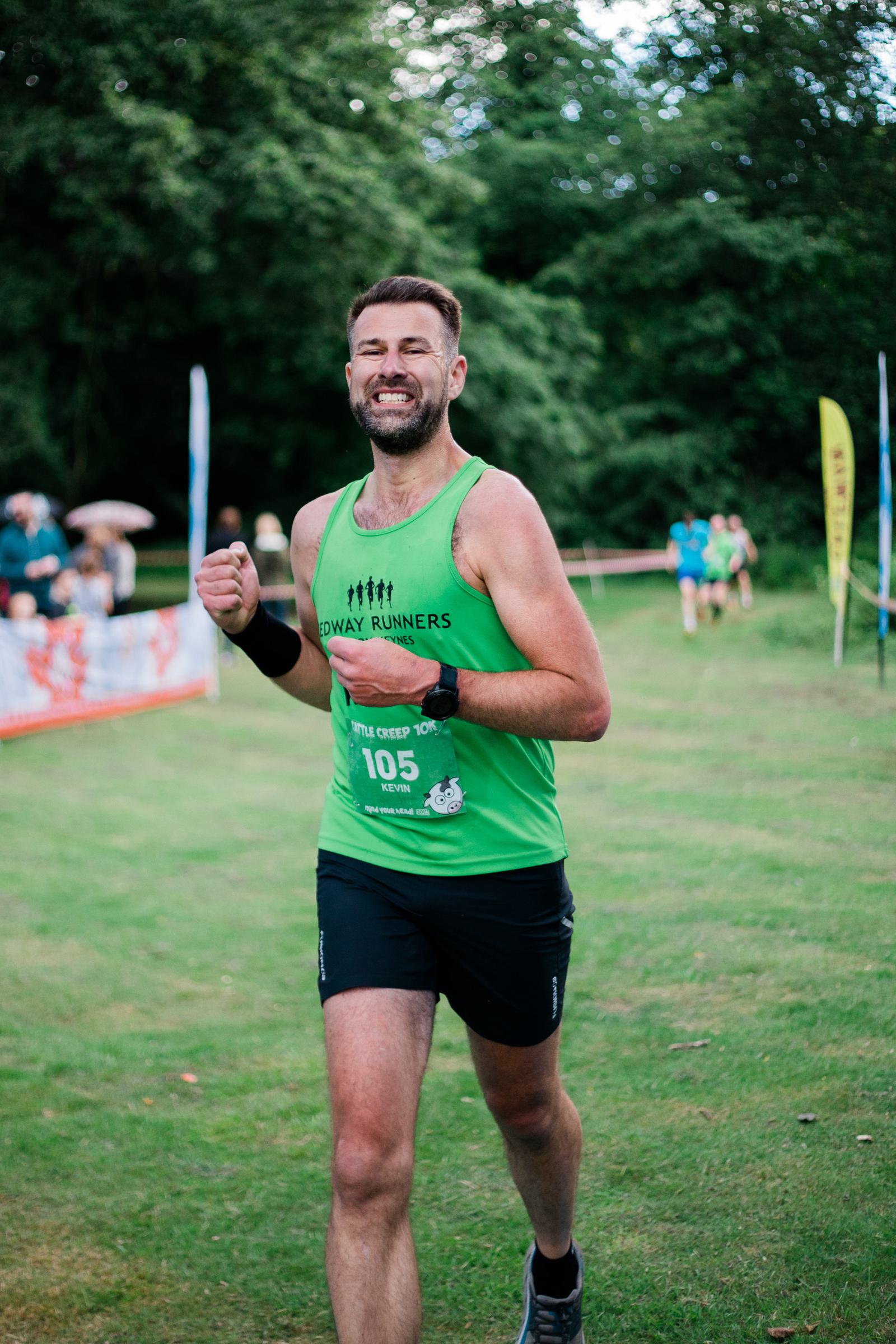 A man in a green tank top and black shorts is running on a grassy field, smiling and giving a thumbs-up. There are trees in the background and a few people watching. He's wearing a race bib numbered "105.