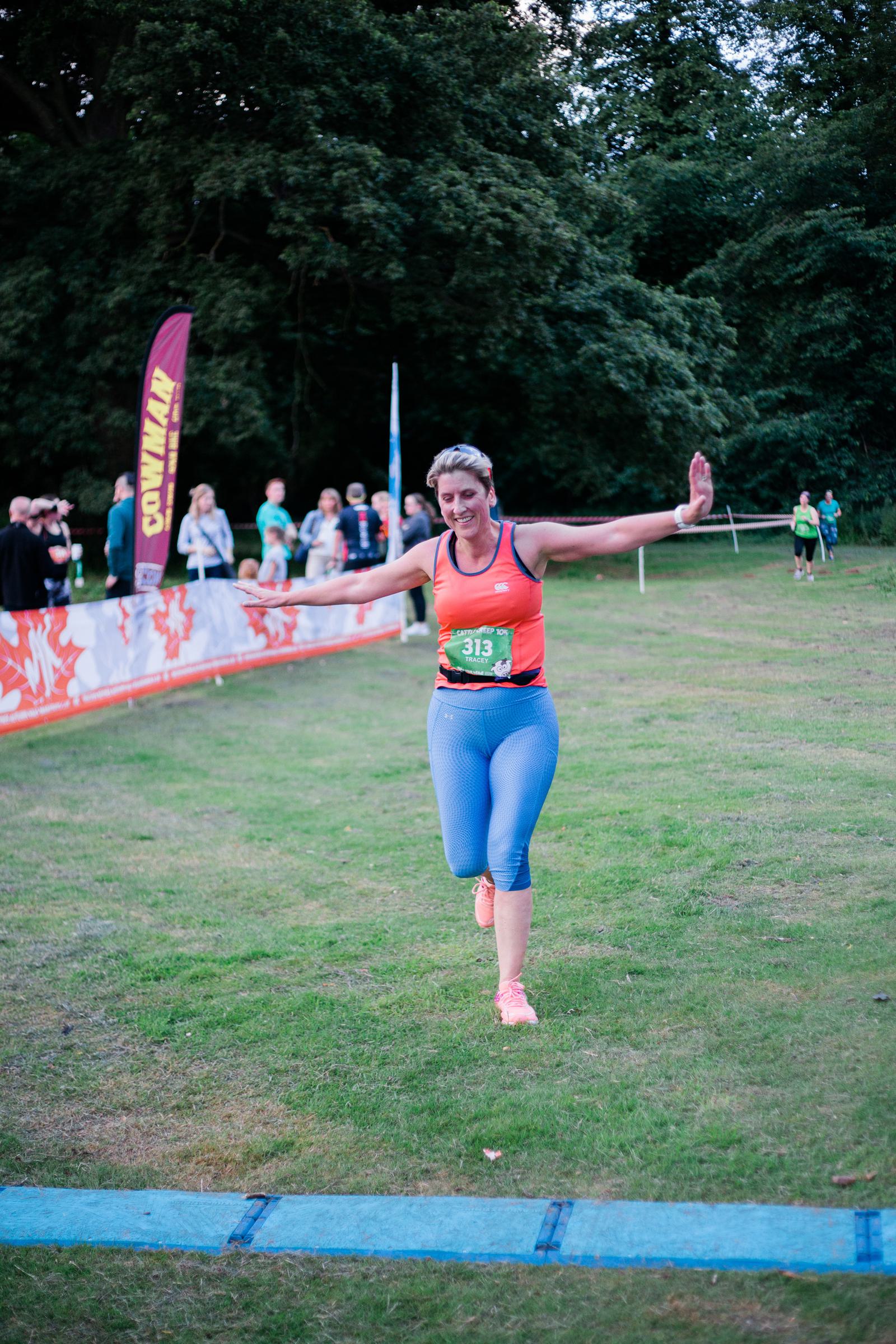 A person in athletic wear joyfully crosses a finish line on a grassy field during a race. They have an arm extended and wear a race bib numbered 213. Spectators and event banners are visible in the background.