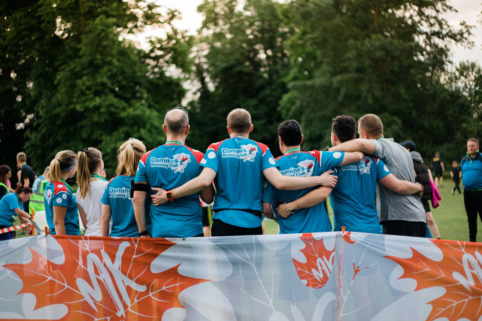 A group of people wearing blue sports shirts with labels on the back stand in an outdoor area. They have their arms around each other, facing away from the camera. A barrier with red and white designs is in the foreground, and trees are in the background.