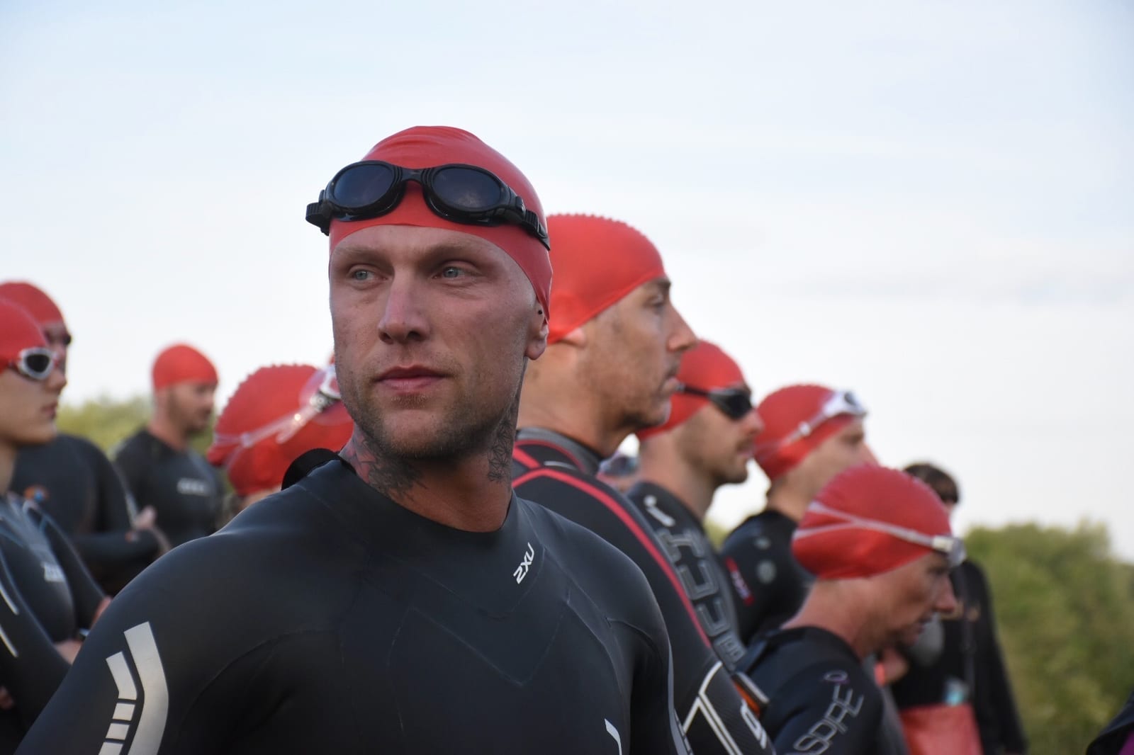 A group of athletes wearing black wetsuits and red swim caps and goggles stand together, preparing for a swimming event. The focus is on the man in the foreground who faces slightly to the left, with other participants blurred in the background.