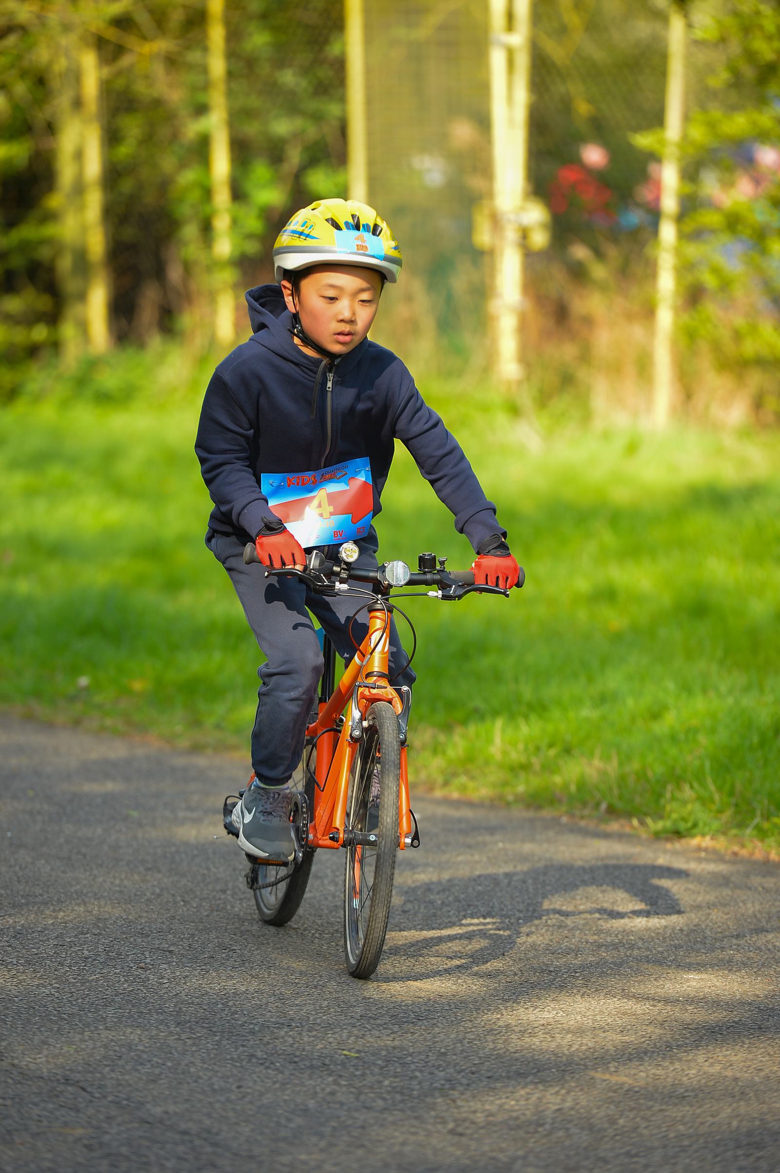 A young child wearing a yellow helmet, blue jacket, and red gloves rides an orange bicycle in a park. A printed number is attached to the front of the bike, suggesting participation in a race or cycling event. Green grass and a fence are visible in the background.