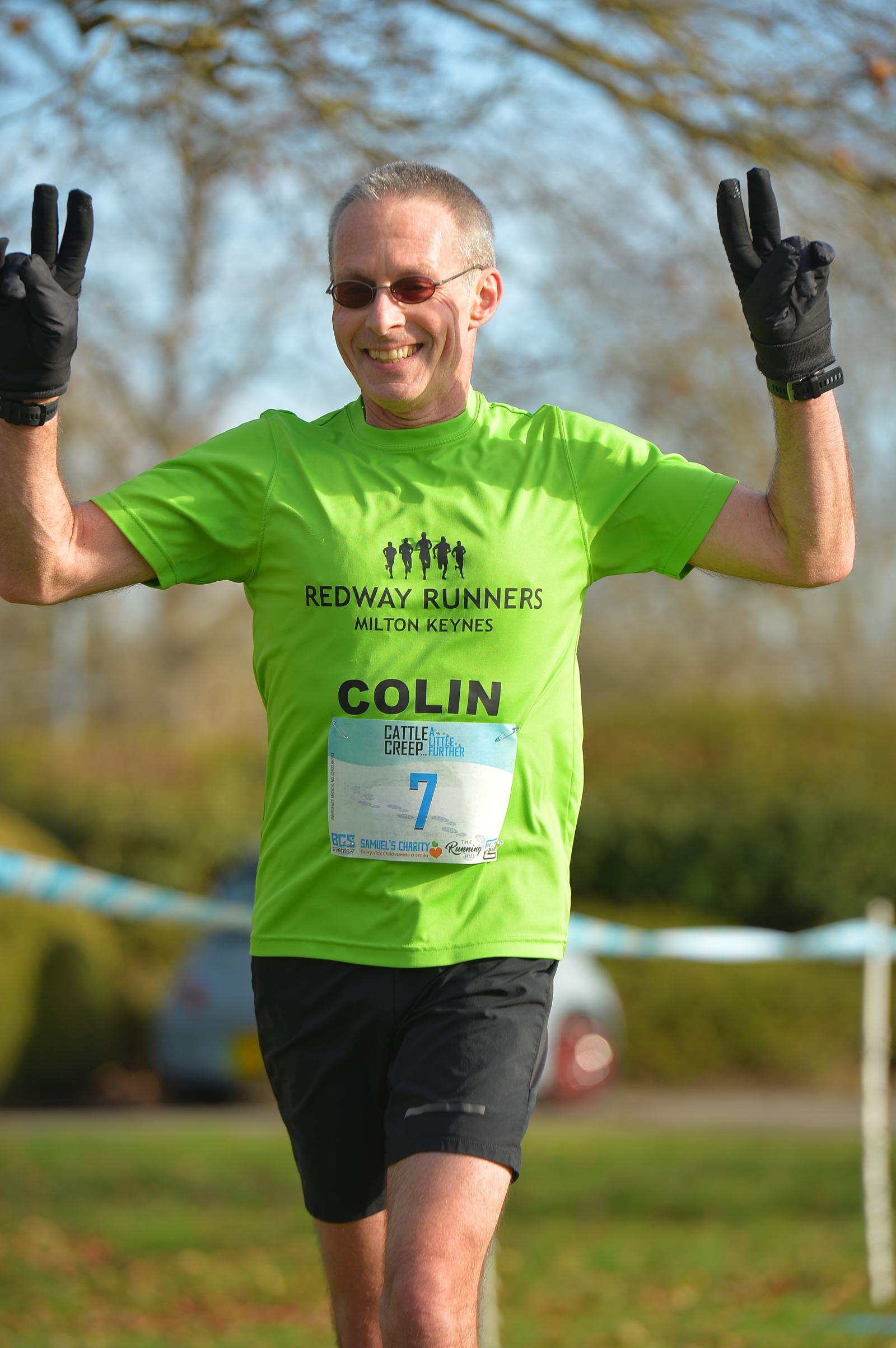 A man in sunglasses and a bright green "Redway Runners Milton Keynes" shirt smiles and raises his arms mid-run. He's wearing black gloves and has a race bib labeled "Colin 7" attached to his shirt. The background is blurred greenery.