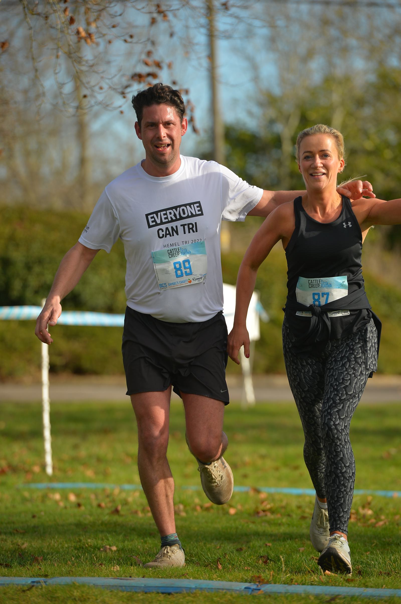 Two people are running a race together, smiling and holding hands. The man wears a white shirt with "Everyone Can Tri" on it, and the woman is in a black tank top and patterned leggings. They are on a grassy path with trees in the background.