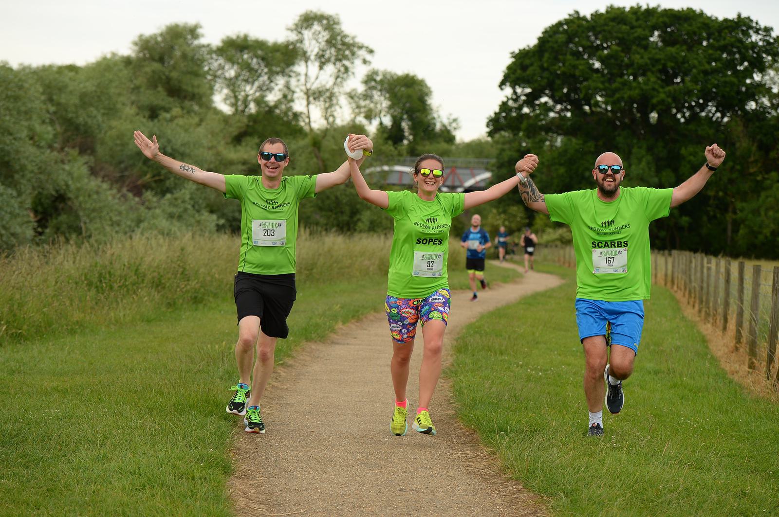 Three smiling runners in green shirts hold hands and celebrate while jogging along a park pathway. They are surrounded by grass and trees, with other runners in the background.