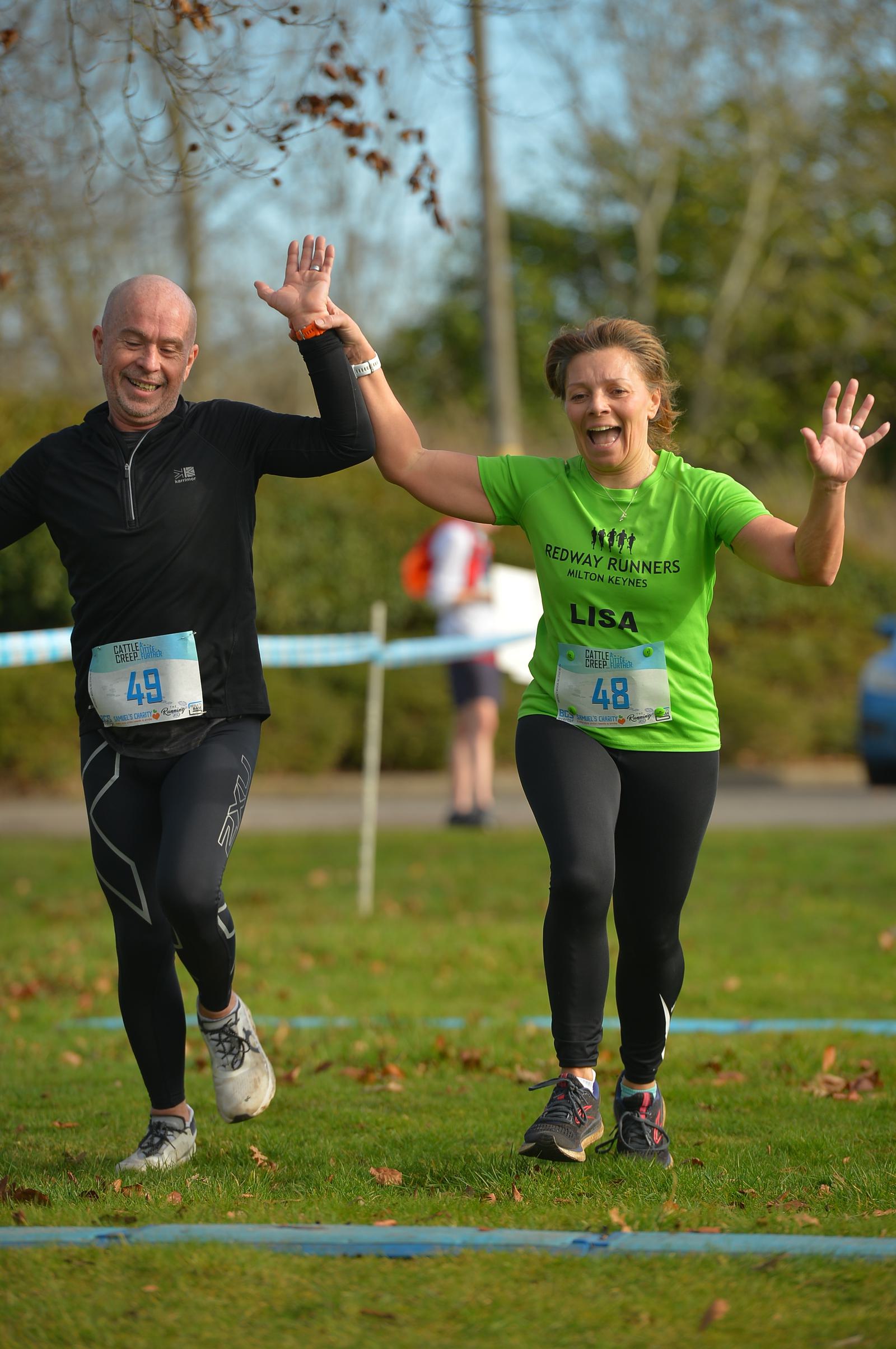 A man and woman happily cross a grassy finish line during a fun run. The man wears a black outfit with a race number 49, while the woman is in a green shirt with number 48. Both have arms raised in excitement. Trees and a spectator are visible in the background.
