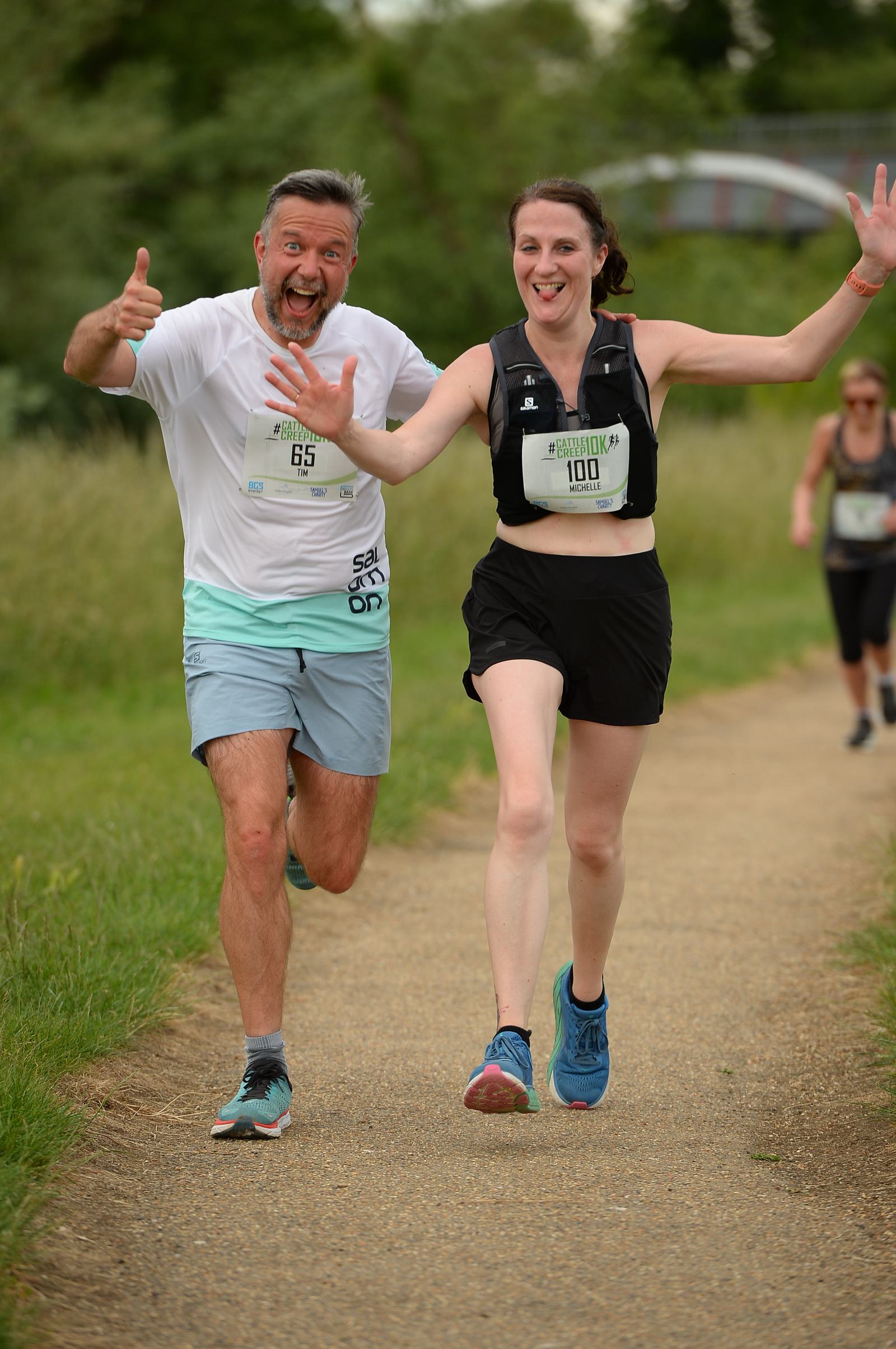 Two smiling runners on a park pathway, giving thumbs up. The man on the left is in a white shirt and blue shorts, and the woman on the right is wearing a black tank top and shorts. Both have race numbers pinned to their clothes.