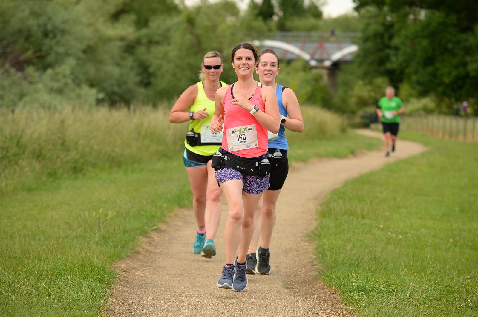 Three women are jogging on a park path, smiling and wearing athletic gear. The runner in the front wears a pink tank top and patterned shorts. Trees and grass surround the path, with a bridge visible in the background.