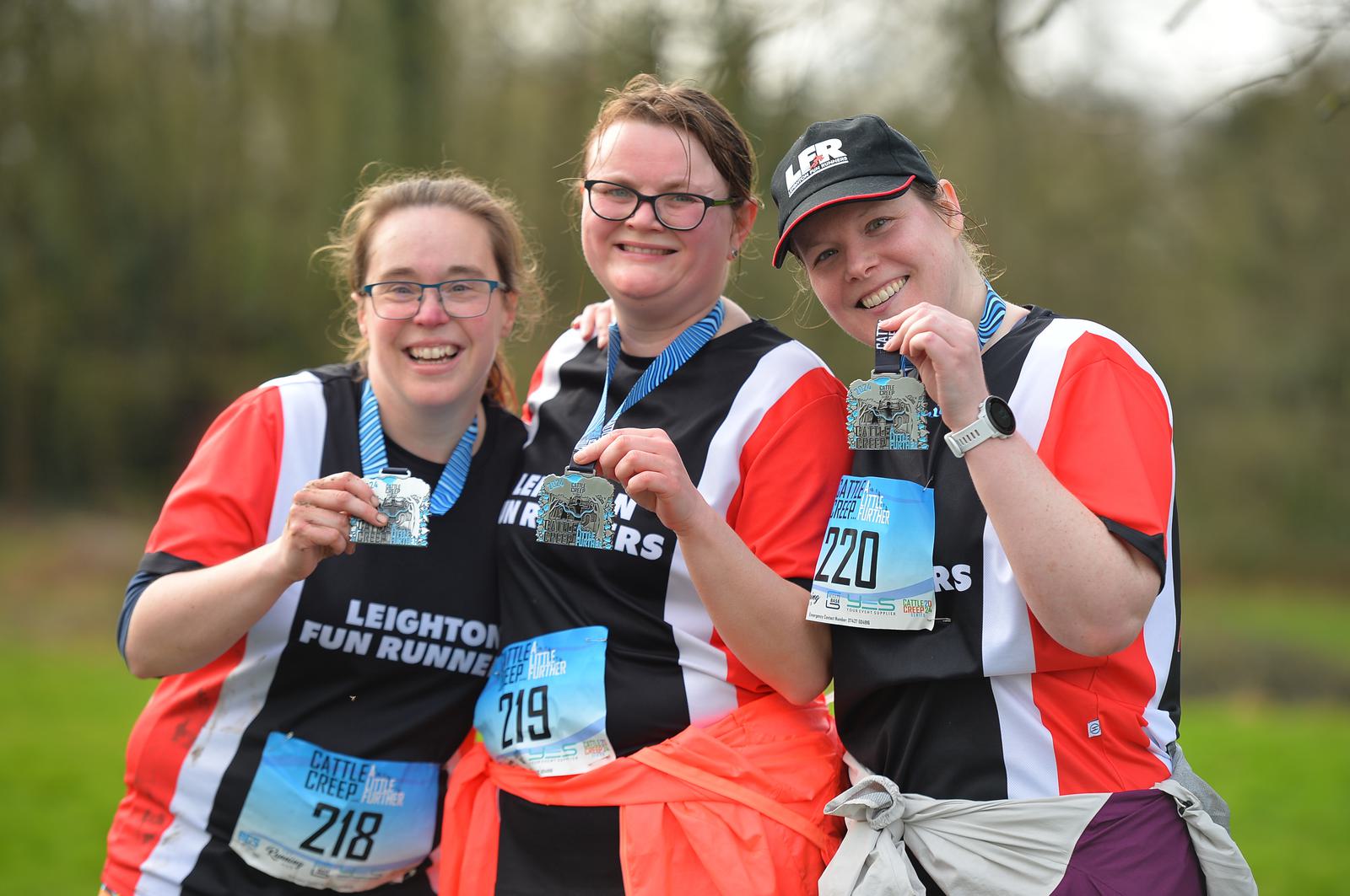 Three smiling women pose together holding medals. They are wearing matching red, black, and white jerseys that read "Leighton Fun Runners," with race bibs numbered 218, 219, and 220. They appear to be outdoors after a running event.