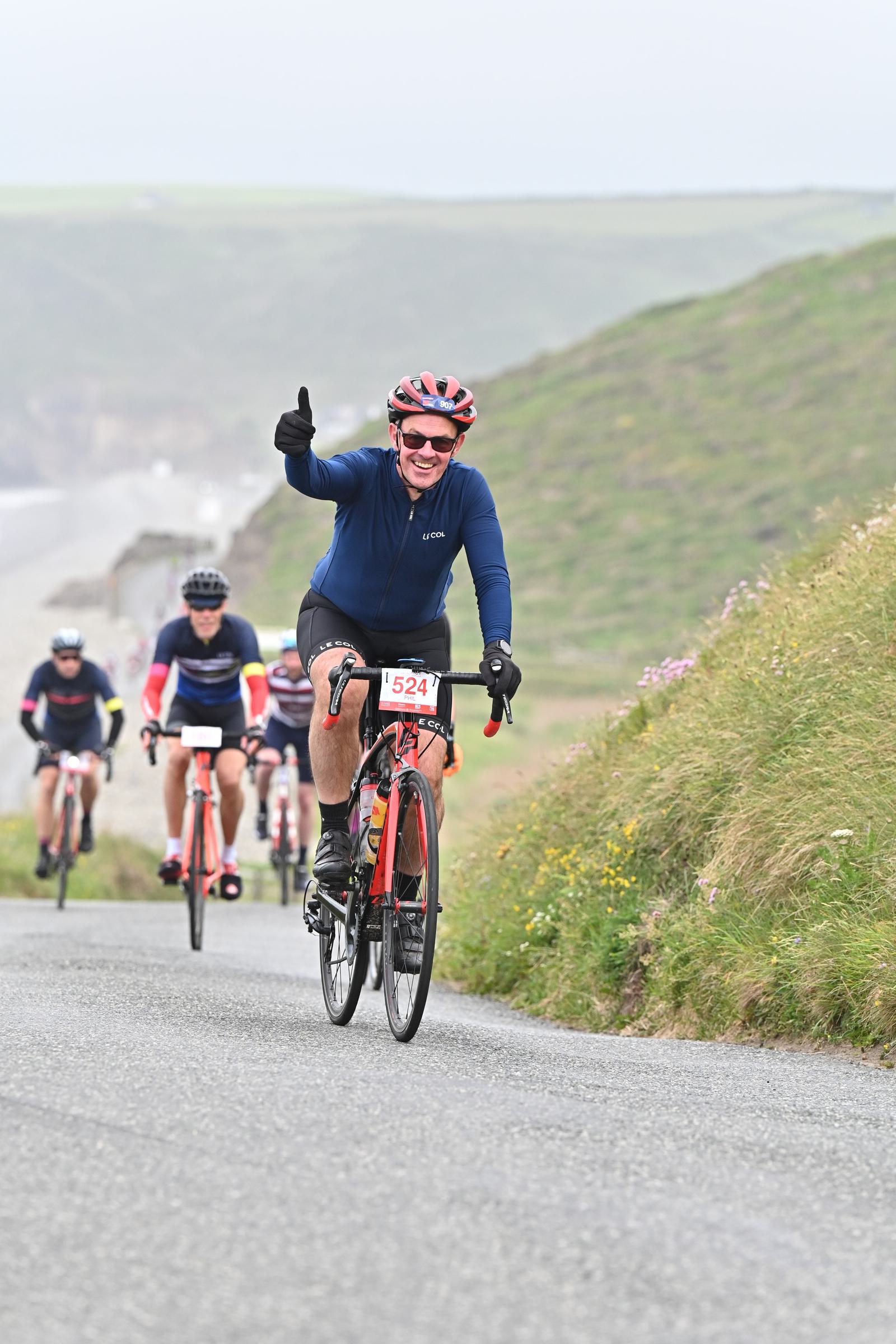 A cyclist wearing a helmet and sunglasses is riding up a hill, smiling and giving a thumbs-up. Several other cyclists follow behind on the rural road lined with grass and wildflowers. The sky is overcast.