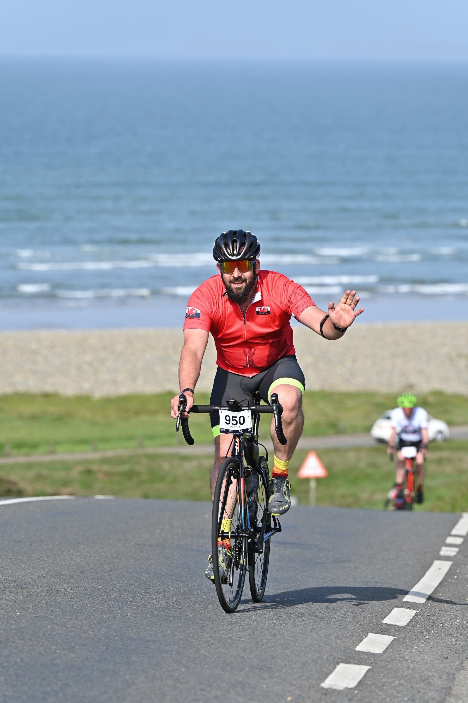 A cyclist in a red jersey waves while riding uphill on a coastal road. The sea and a green landscape are in the background. Another cyclist is seen further back. The cyclist is wearing a helmet and sunglasses.