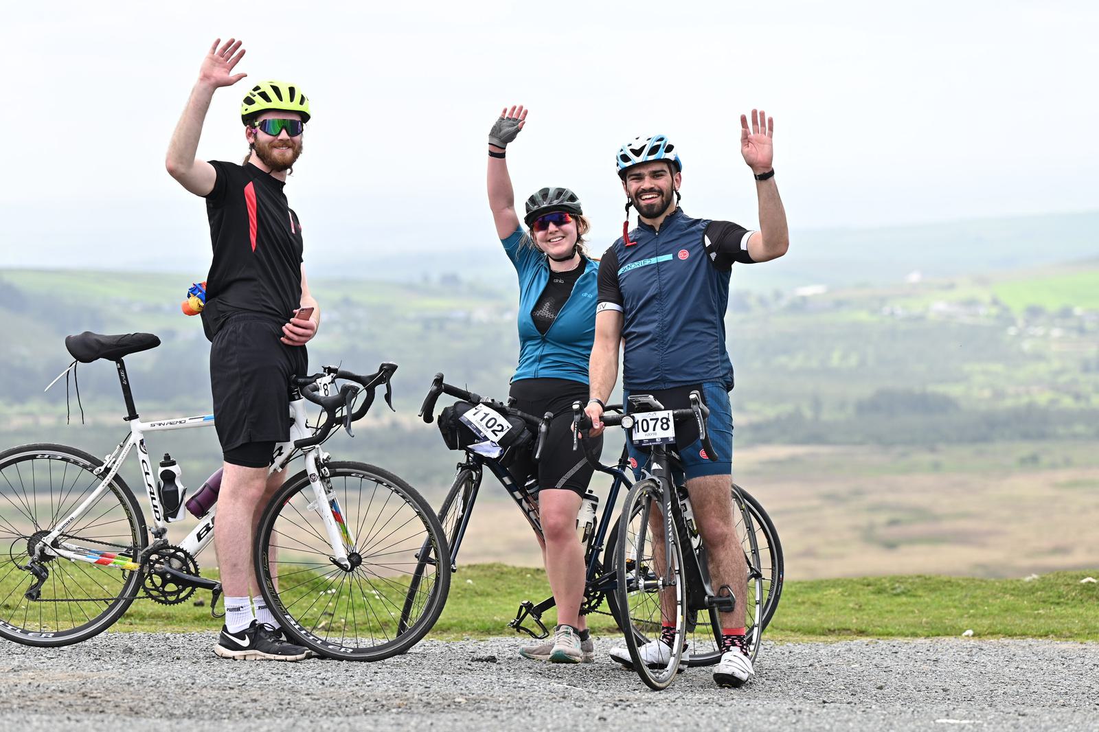 Three cyclists wearing helmets and sports gear pause to wave at the camera. They are standing with their bikes on a hill overlooking a scenic landscape. Two men and a woman are smiling, dressed in casual cycling attire.
