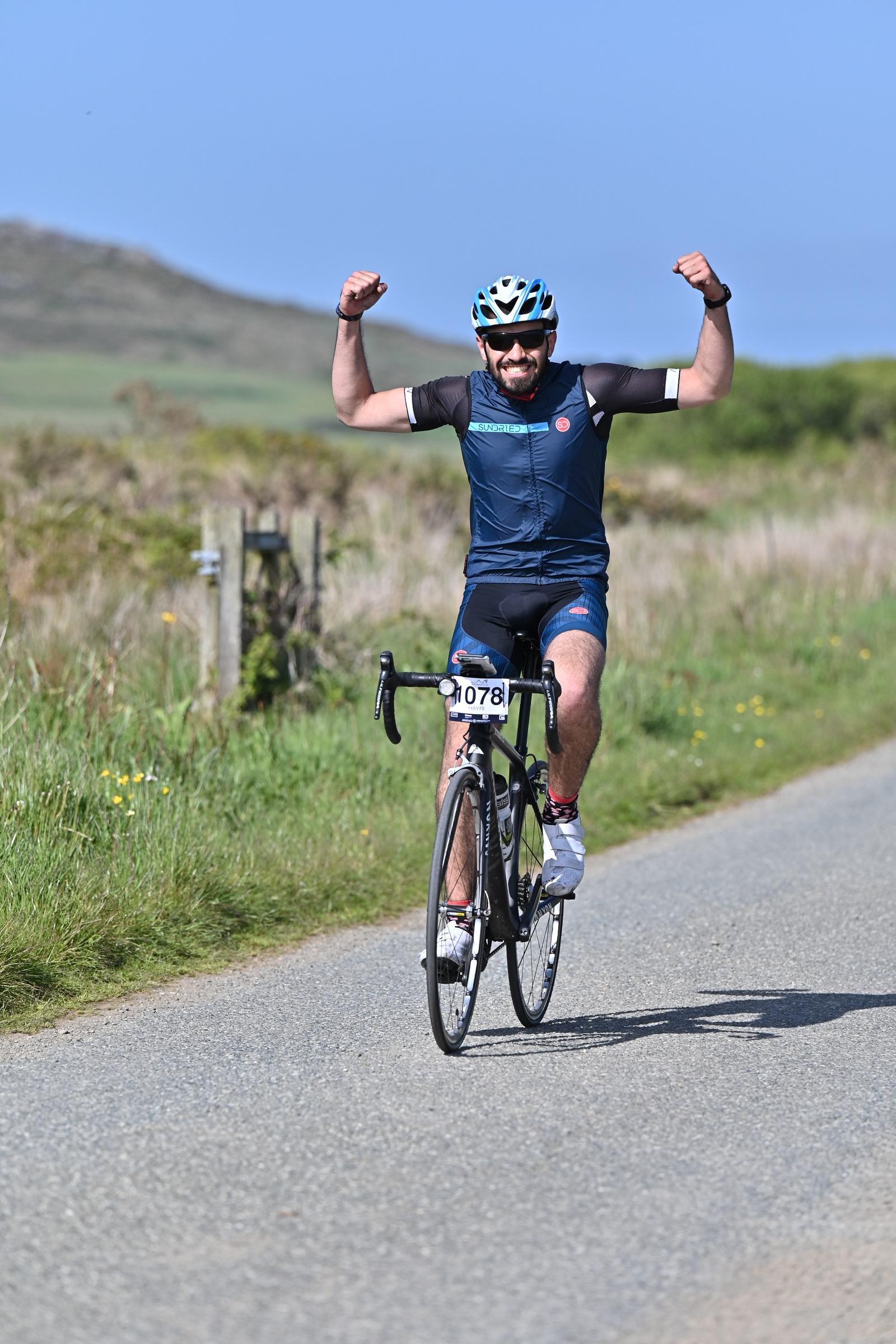 A cyclist in a blue outfit and helmet celebrates with raised arms while riding a bicycle on a rural road. A race number is visible on the bike. The background features grassy fields and a hill under a clear blue sky.