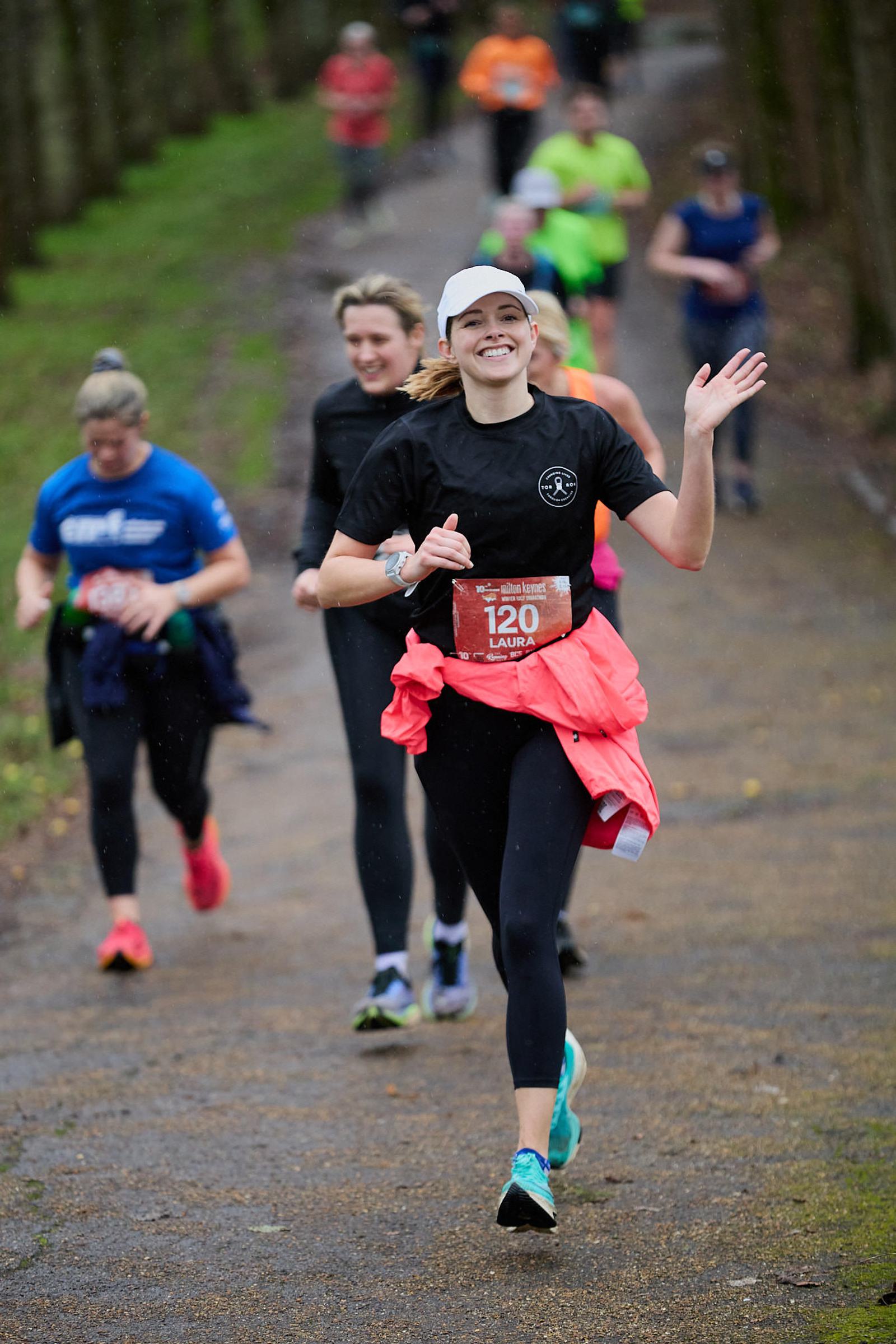 A woman in athletic wear, smiling and waving, jogs on a park pathway during a race. She wears a white cap and a red race bib with the number 120. Other runners are behind her, and trees line the path.
