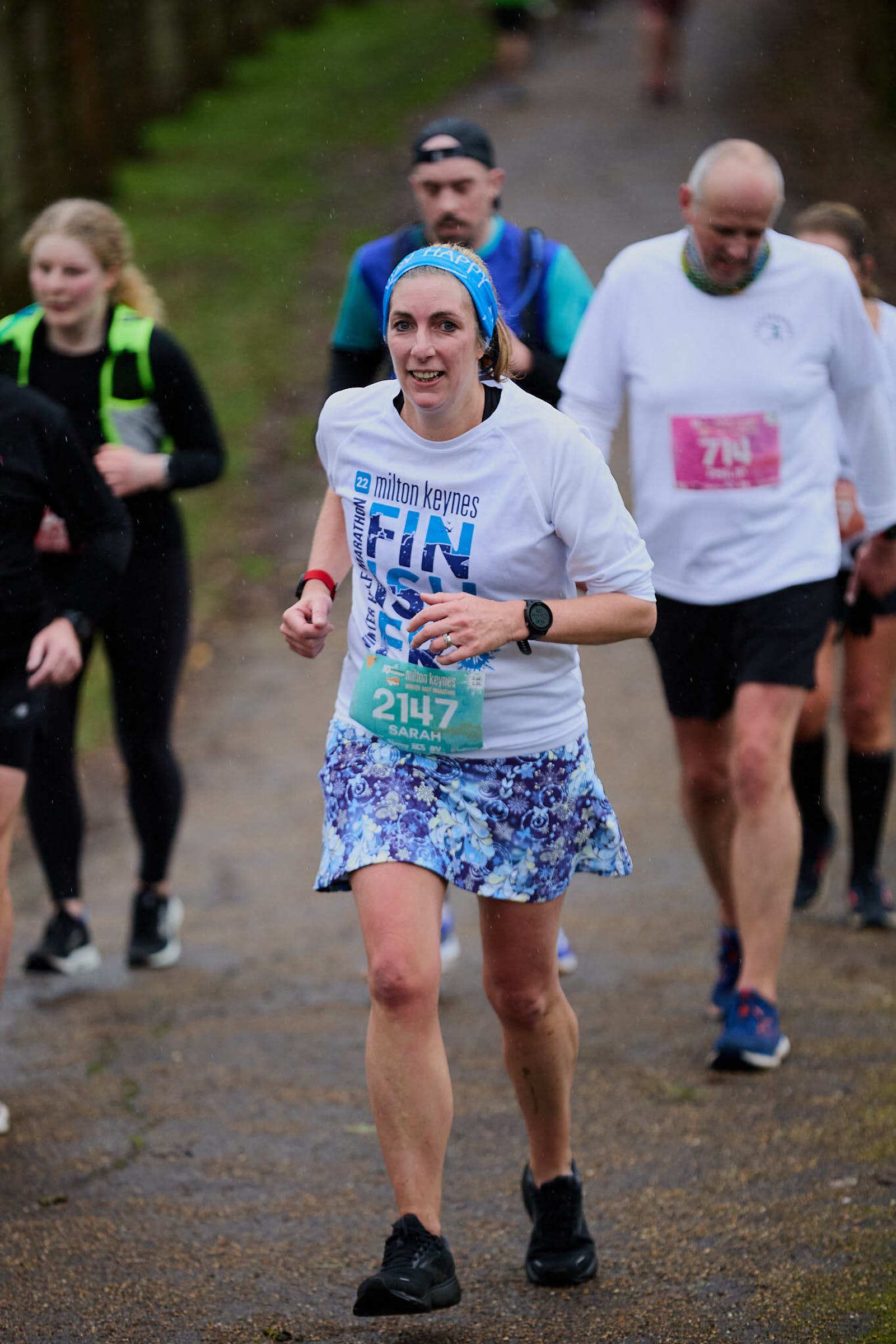 A woman wearing a blue headband, white shirt, and patterned skirt runs on a wet road during a race. Other runners are visible in the background. It appears to be a cloudy day.