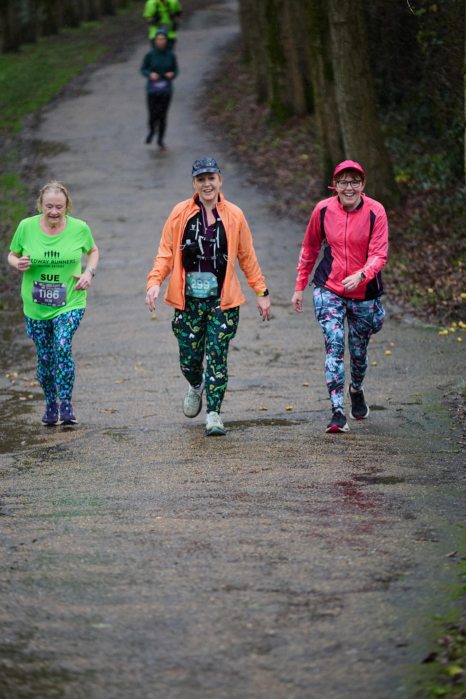 Three women with colorful outfits walk together on a wet, wooded path during a race. One wears a green shirt, another an orange jacket, and the third a red cap. They are smiling and appear to be enjoying the event.