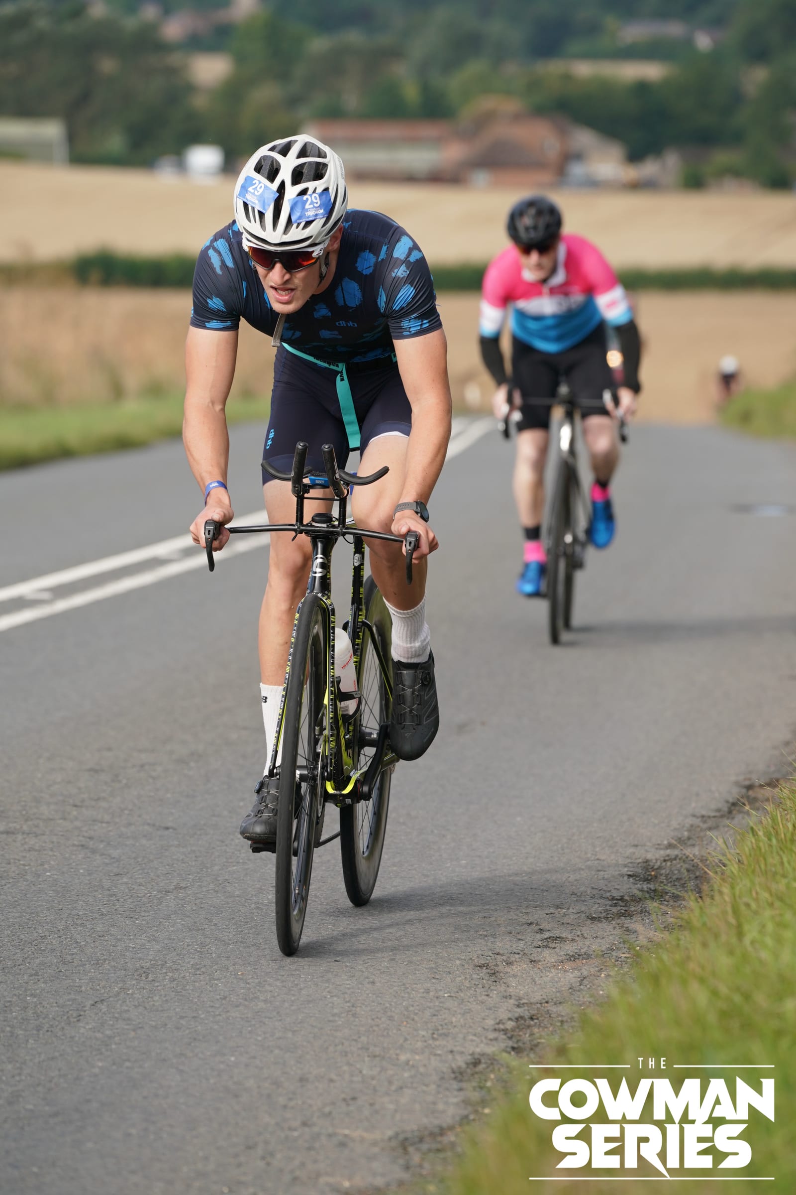 Two cyclists ride uphill on a rural road during a race. The cyclist in front wears a blue helmet and sportswear, while the second cyclist follows behind. "The Cowman Series" logo is visible in the lower right corner.