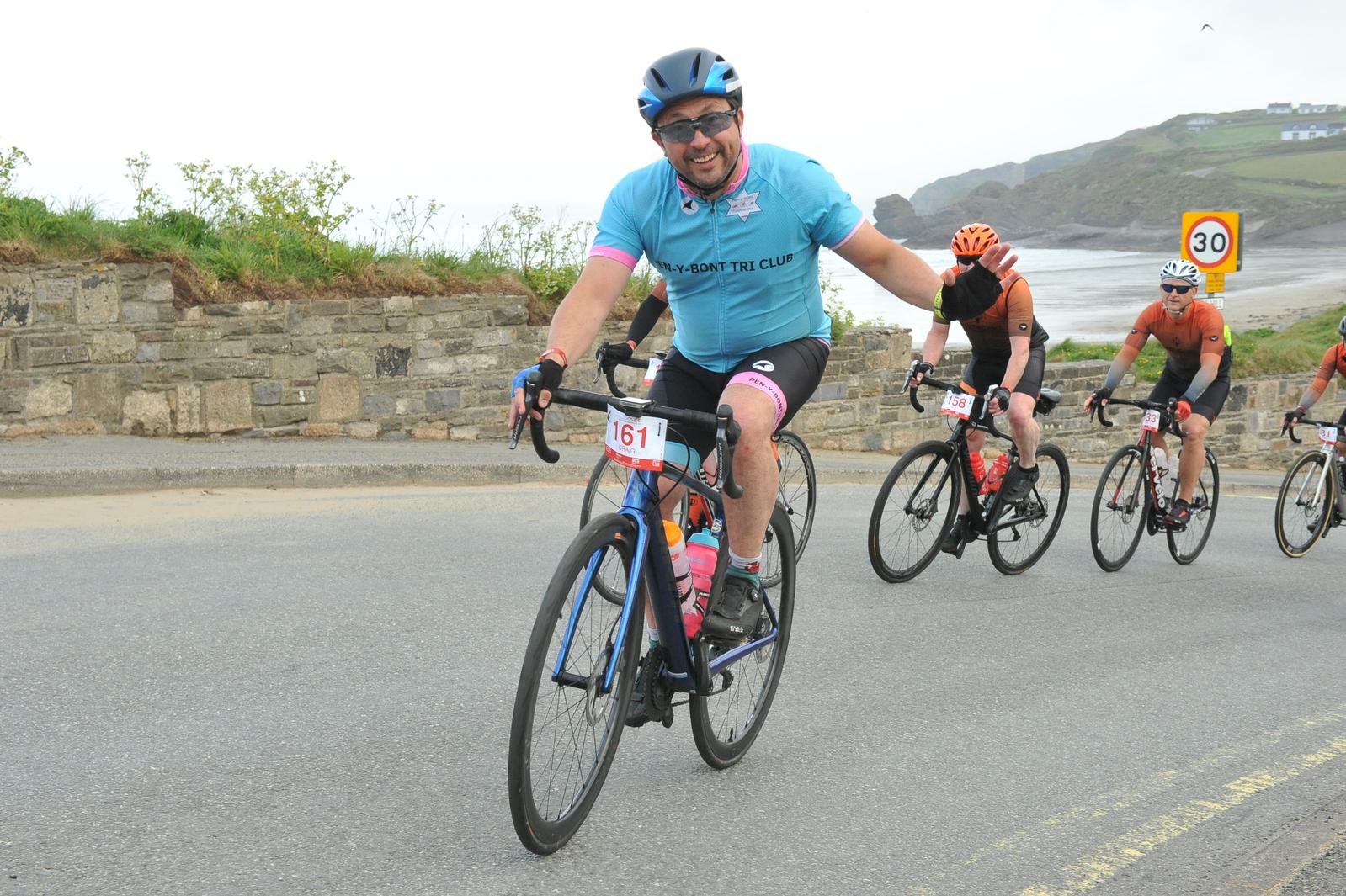 A cyclist in a blue jersey and helmet waves and smiles while riding on a road near the coast. Other cyclists follow behind him. The background features a rocky shoreline and greenery. There's a speed limit sign visible on the roadside.