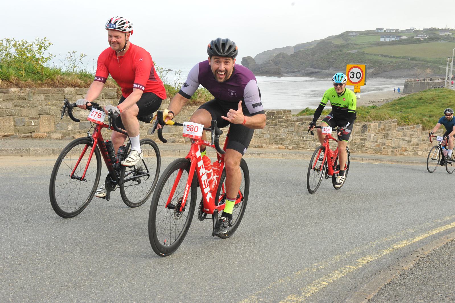 Three cyclists ride on a coastal road. The cyclist in the center wearing purple smiles and gestures at the camera. The landscape includes hills, a beach, and a road sign indicating 30 mph. All cyclists wear helmets and numbered bibs.