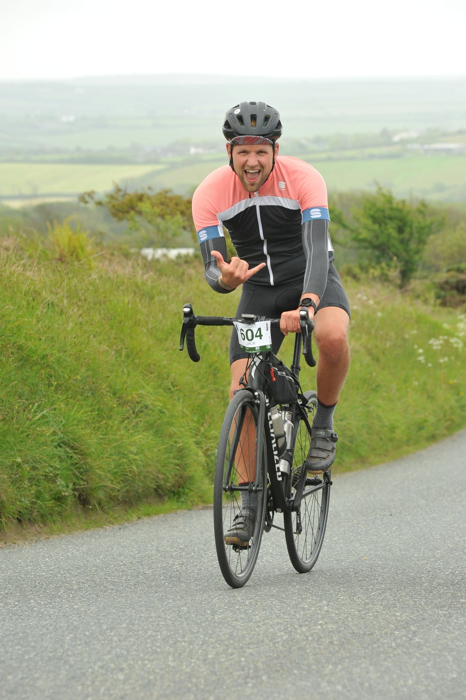 A cyclist wearing a pink and black jersey and helmet rides uphill on a country road, joyfully pointing and sticking out their tongue. The landscape features lush green fields and a cloudy sky in the background.