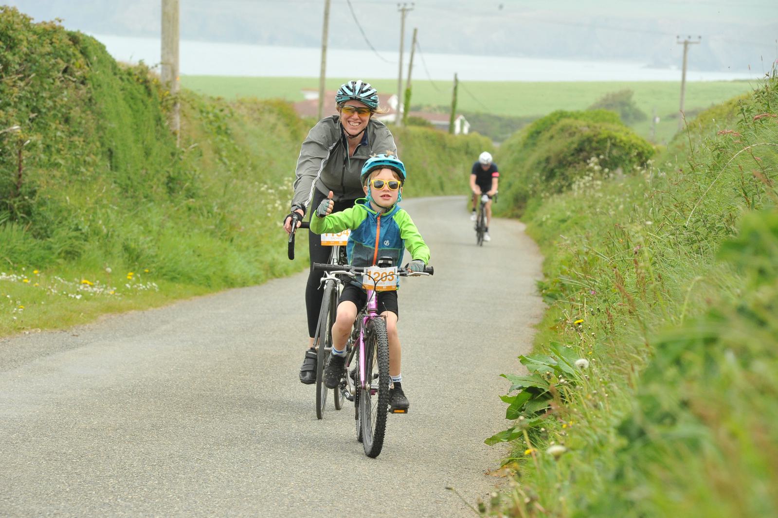 A child wearing a green jacket and blue pants rides a bicycle with number 295, accompanied by an adult in a gray jacket and helmet on a rural road. Lush greenery surrounds them, with another cyclist in the distance.