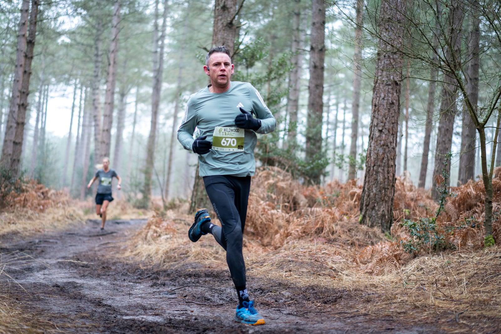 A man wearing athletic gear and a race bib runs on a muddy forest trail, with another runner visible in the background among tall trees and fallen leaves.
