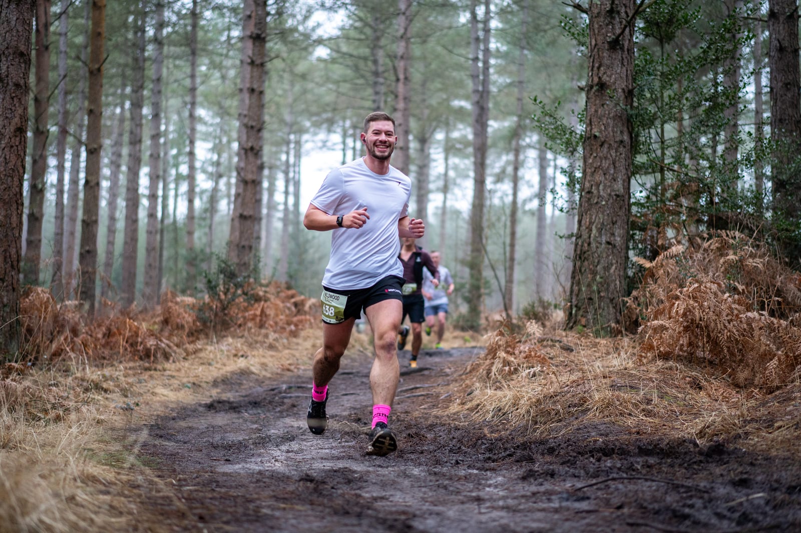 A man in a white shirt and pink socks runs on a muddy forest trail, smiling, with other runners visible in the background among tall trees and brown foliage.
