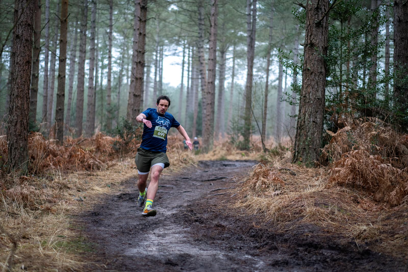 A person in a blue shirt and shorts is running energetically along a muddy forest trail. They are surrounded by tall trees and brown ferns, with a misty background. The path is narrow and lined with rough terrain.