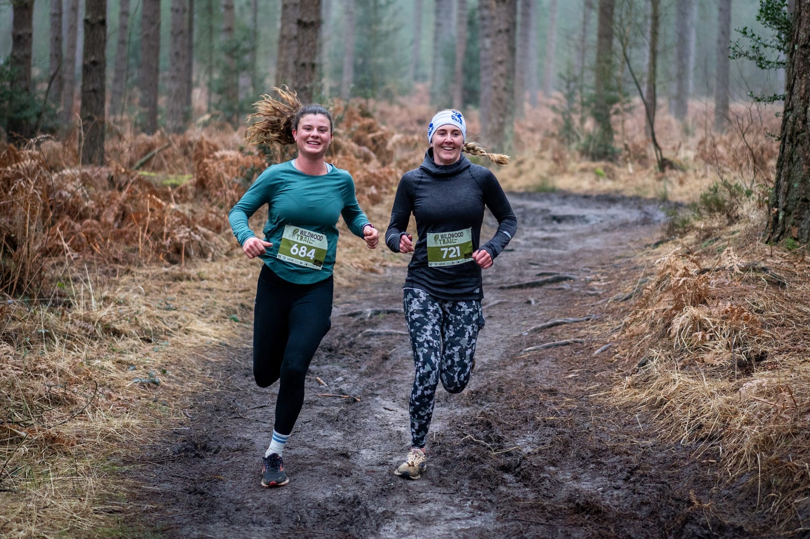 Two women with race bibs run and smile on a muddy forest trail. They wear athletic clothing and appear to be enjoying the outdoor event surrounded by trees and brown foliage.