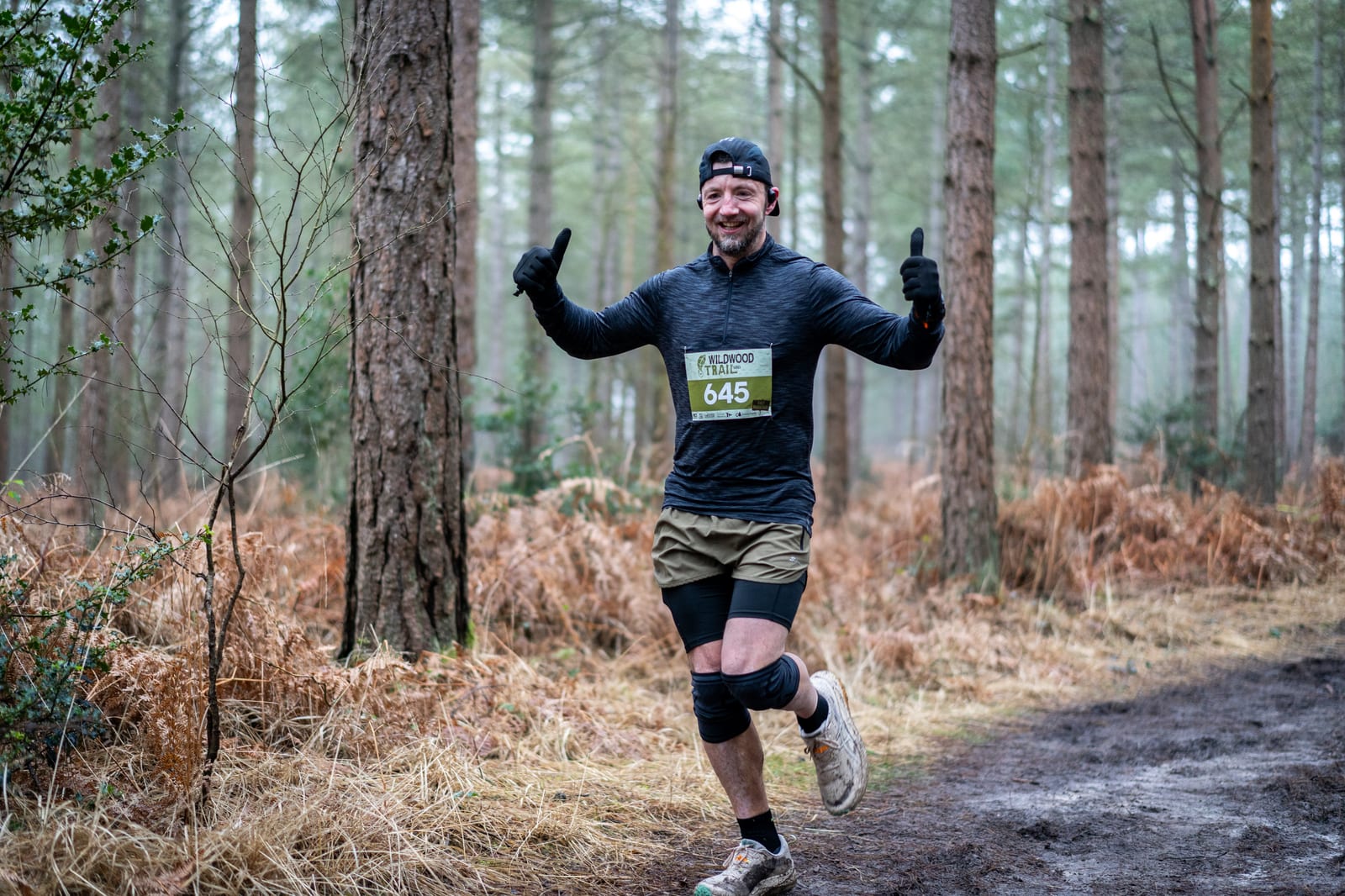 A smiling runner wearing a race bib, black shirt, shorts, and a cap gives two thumbs up while jogging on a forest trail surrounded by tall trees and brown foliage.