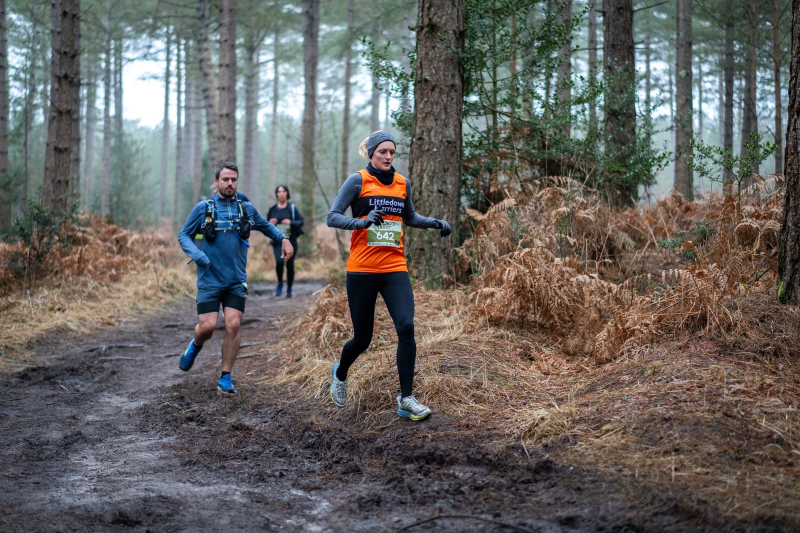 Runners participate in a trail race through a forest. A person in an orange top leads, followed by others. The path is muddy and surrounded by trees and ferns. The atmosphere is cool and misty.