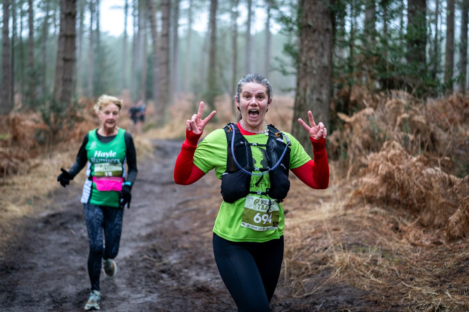 Two women are trail running in a forest. The woman in front is wearing a green shirt and making peace signs with both hands while smiling. The woman in the background wears a green and black top, jogging along the muddy path surrounded by tall trees.