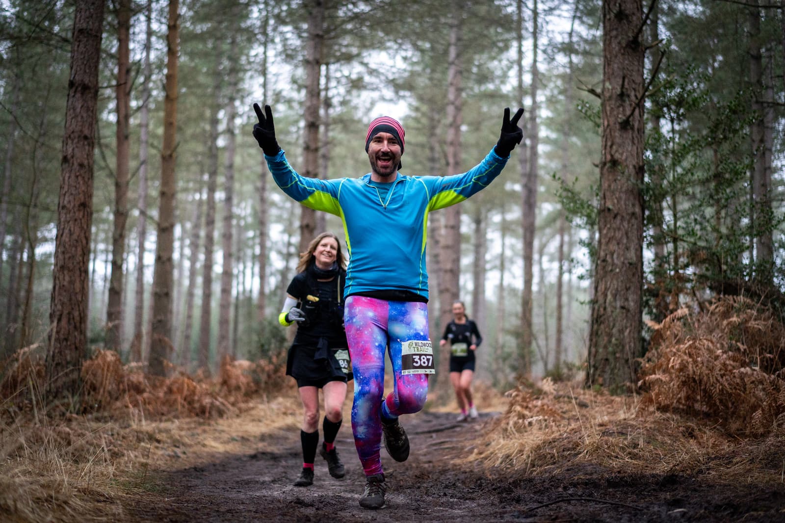 A man in colorful running attire joyfully runs through a forest trail, holding up peace signs with both hands. Two other runners follow behind. The scene is surrounded by tall trees and a misty atmosphere.