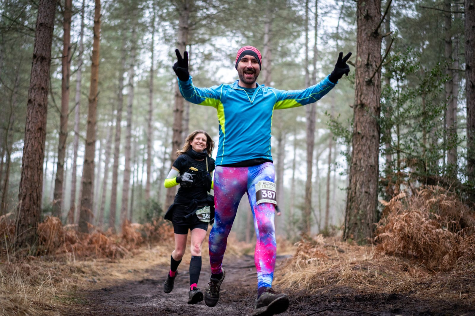 Two runners are on a forest trail. The man in front is wearing colorful leggings and a blue top, smiling and making peace signs with both hands, while the woman behind him smiles as she runs. Both are wearing race bibs.