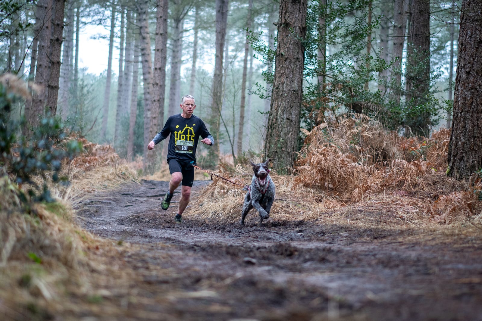 A man in athletic gear runs on a muddy forest trail with a dog on a leash. Tall trees and brown brush line the path, and the scene appears misty and cool.