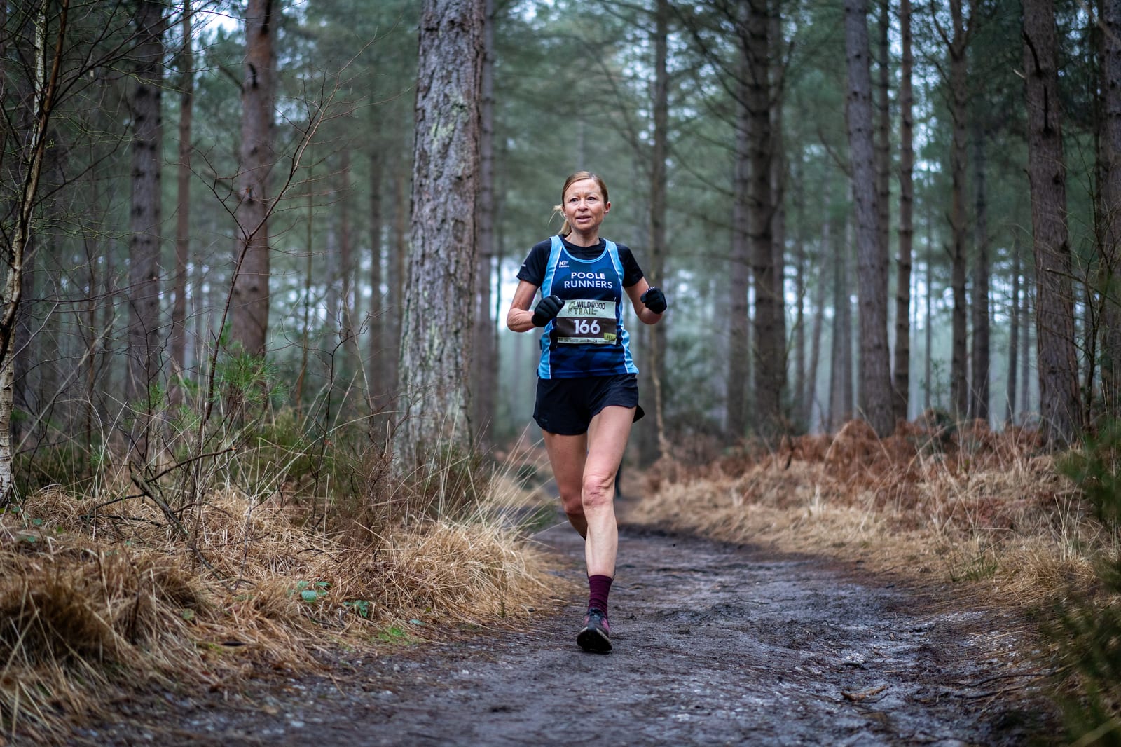 A woman in athletic gear runs on a muddy forest trail surrounded by tall trees and dry grass, wearing a race bib and looking ahead.