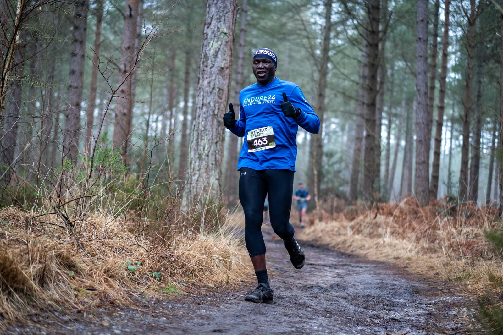 A person is running on a forest trail, smiling and giving a thumbs-up. They are wearing a blue long-sleeve shirt, black leggings, and a headband. The path is surrounded by tall, sparse trees and dry grass. It's a misty day.