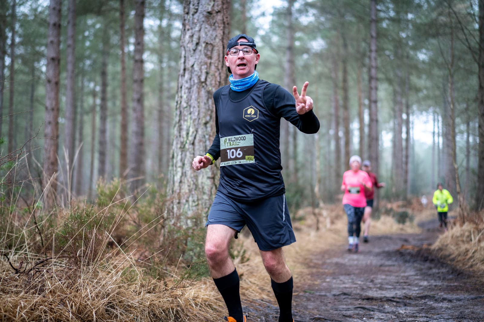 A man in athletic gear is running on a forest trail, holding up a peace sign with one hand. He wears a headband, glasses, and a race number on his chest. Other runners are blurred in the background among tall trees. The scene is overcast.