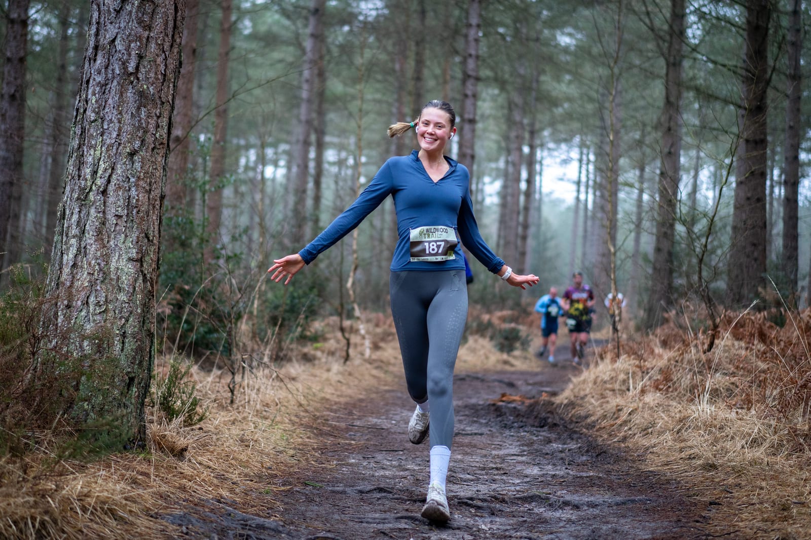 A woman in athletic clothing, wearing bib number 187, runs joyfully along a muddy forest trail. Trees and dry grass surround her, and other runners are visible in the background.