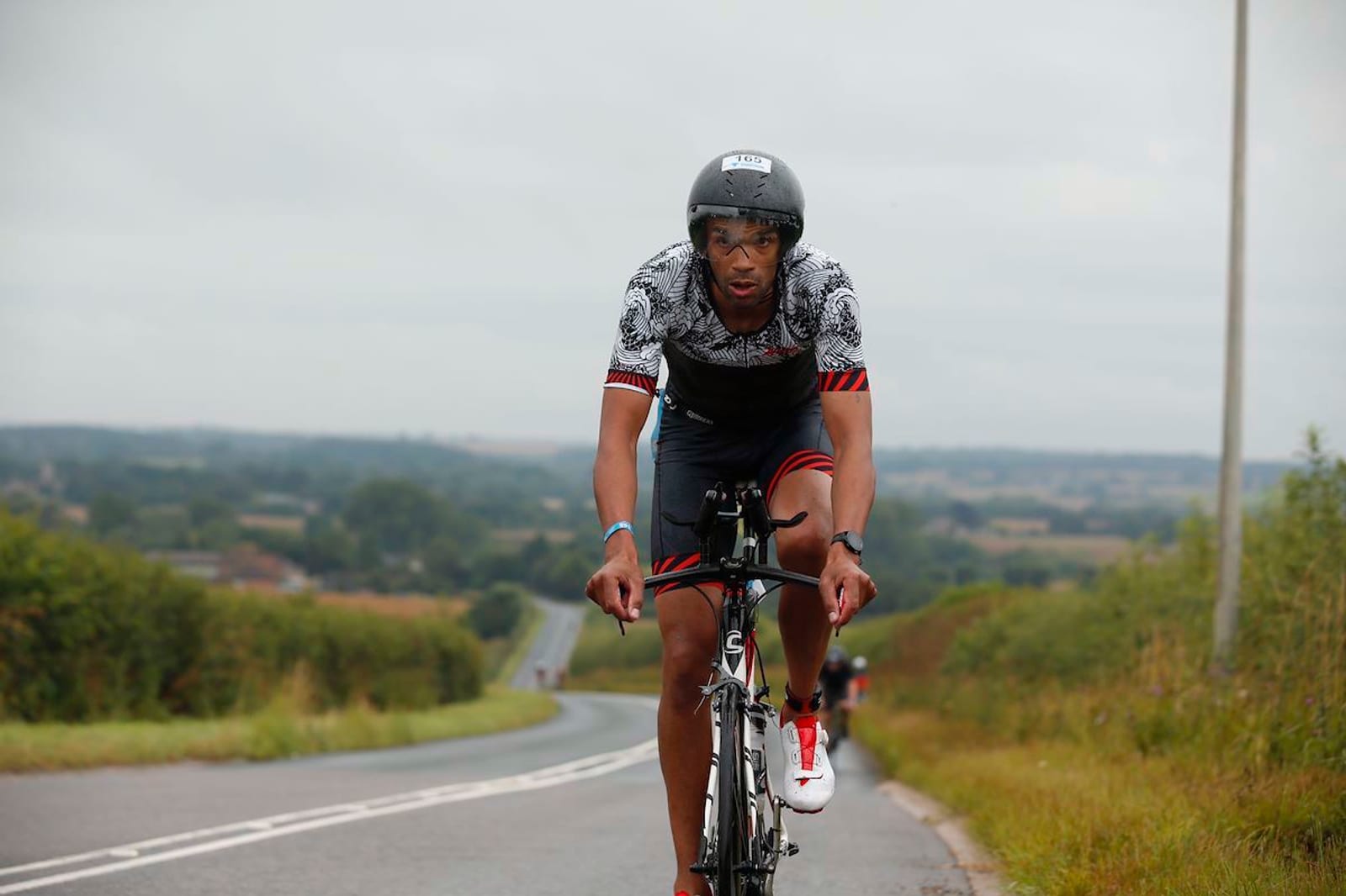 A cyclist wearing a helmet, sunglasses, and a patterned outfit rides a bicycle on a paved road through a rural landscape. The sky is overcast, and the background features fields and distant trees.