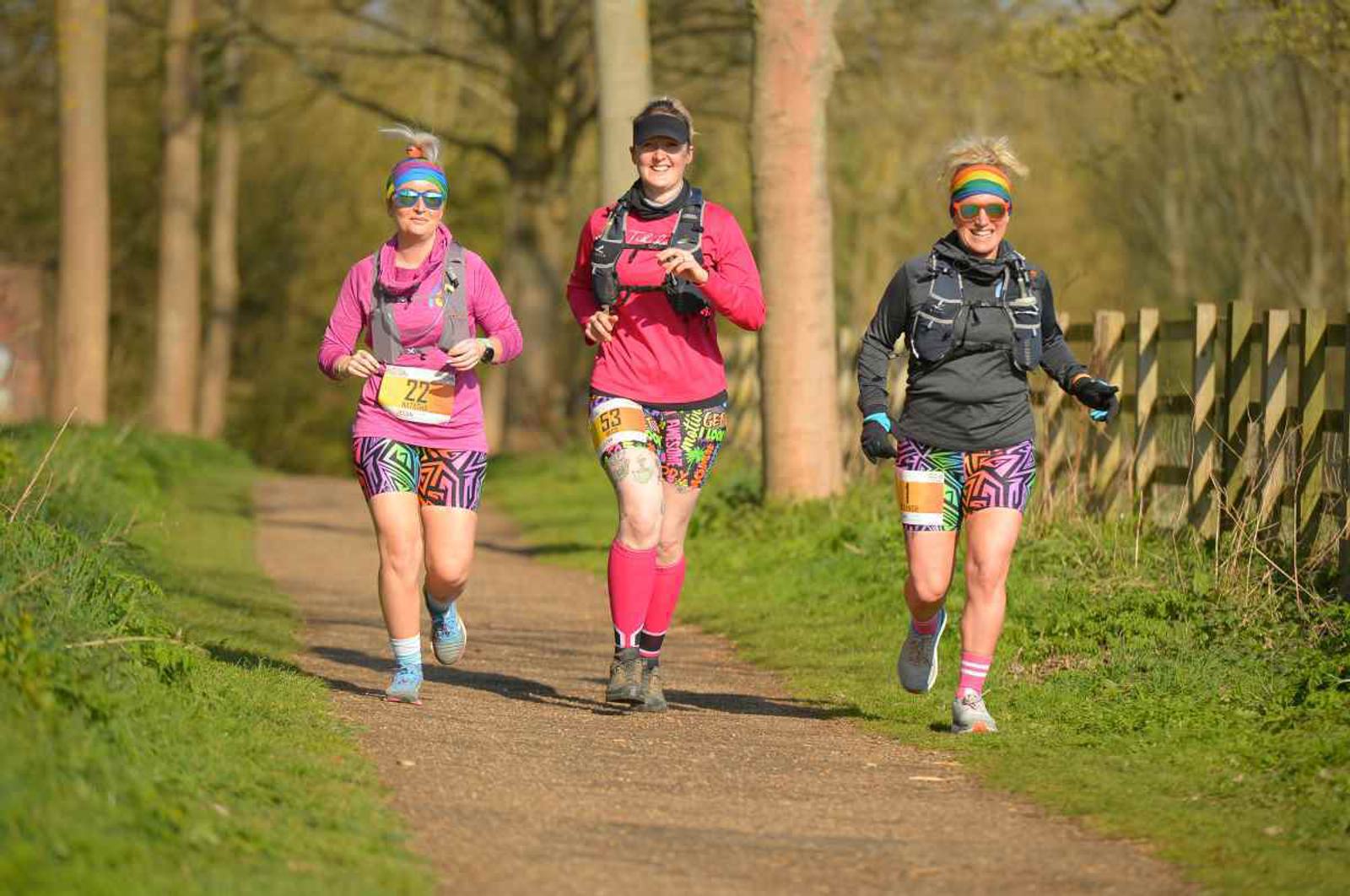 Three people jog along a sunny trail, wearing colorful athletic outfits and numbered race bibs. They're surrounded by trees and grass, exuding a cheerful, energetic vibe as they participate in an outdoor event.