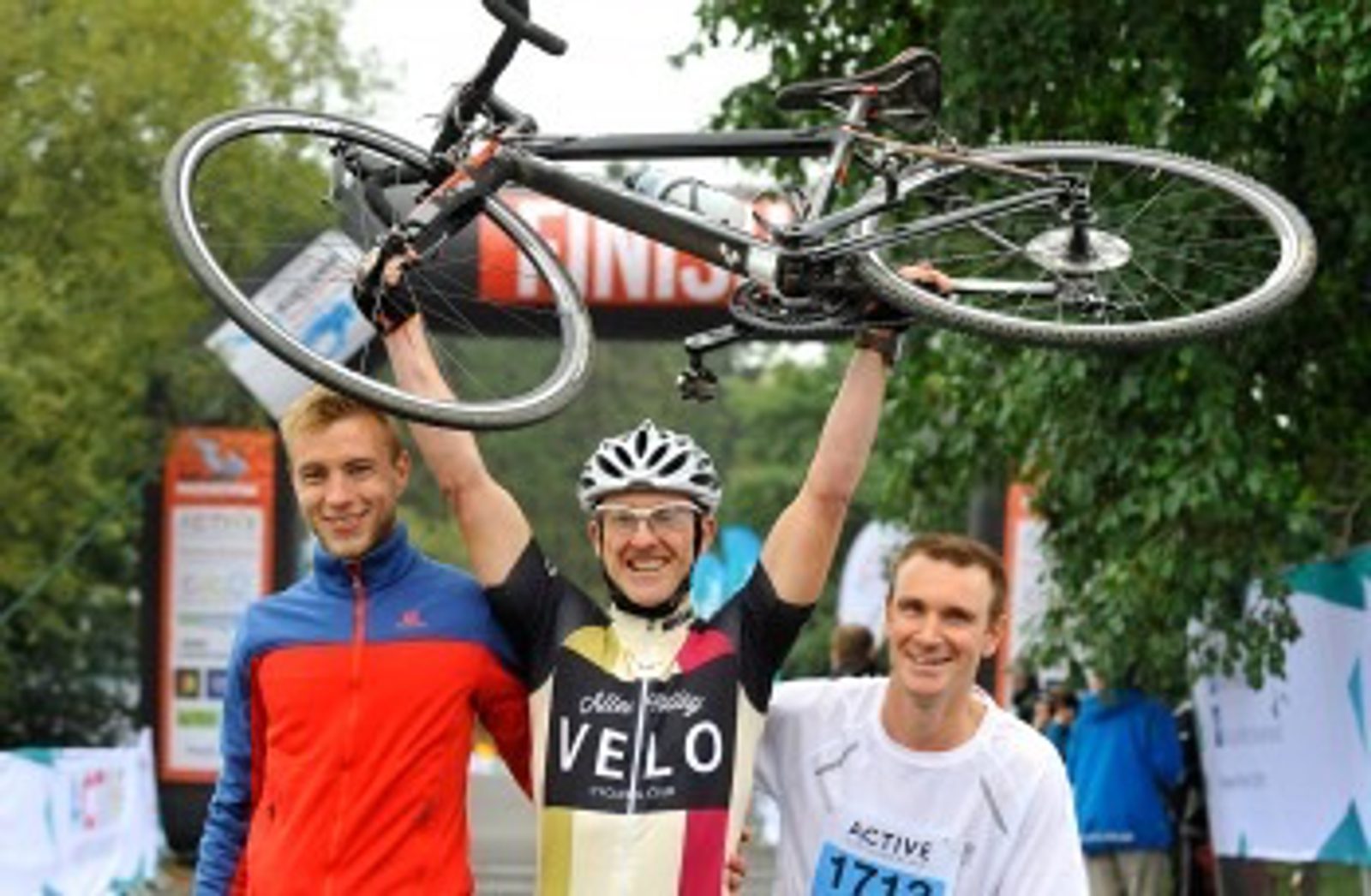 Three men celebrate at a race finish line. The man in the center triumphantly holds a bicycle over his head while wearing cycling gear. The other two men stand on either side, smiling and wearing athletic apparel. A "FINISH" sign is visible in the background.