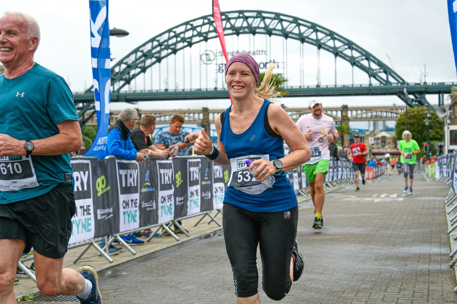 Runners participating in a race pass near the Tyne Bridge. A woman in blue and black athletic wear with bib number '53' smiles as she runs on a paved path lined with event barriers. Other participants and onlookers are visible in the background.