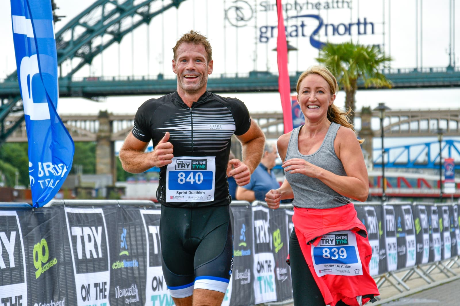 Two athletes, a man and a woman, smile and run side by side wearing race bibs numbered 840 and 839, respectively. They are participating in a race, with the background showing part of a large bridge and event banners. The woman has a red jacket tied around her waist.