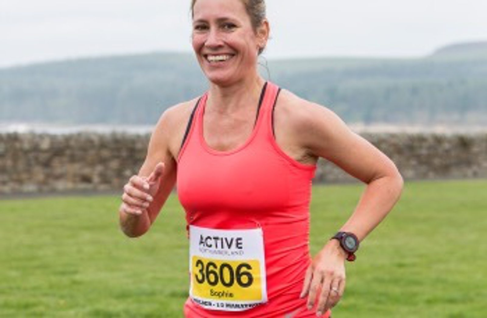 A woman is running outdoors wearing a bright pink tank top and a race bib number 3606. She is smiling and appears to be in mid-stride. Behind her is a grassy area, a low stone wall, and distant hills under a cloudy sky.